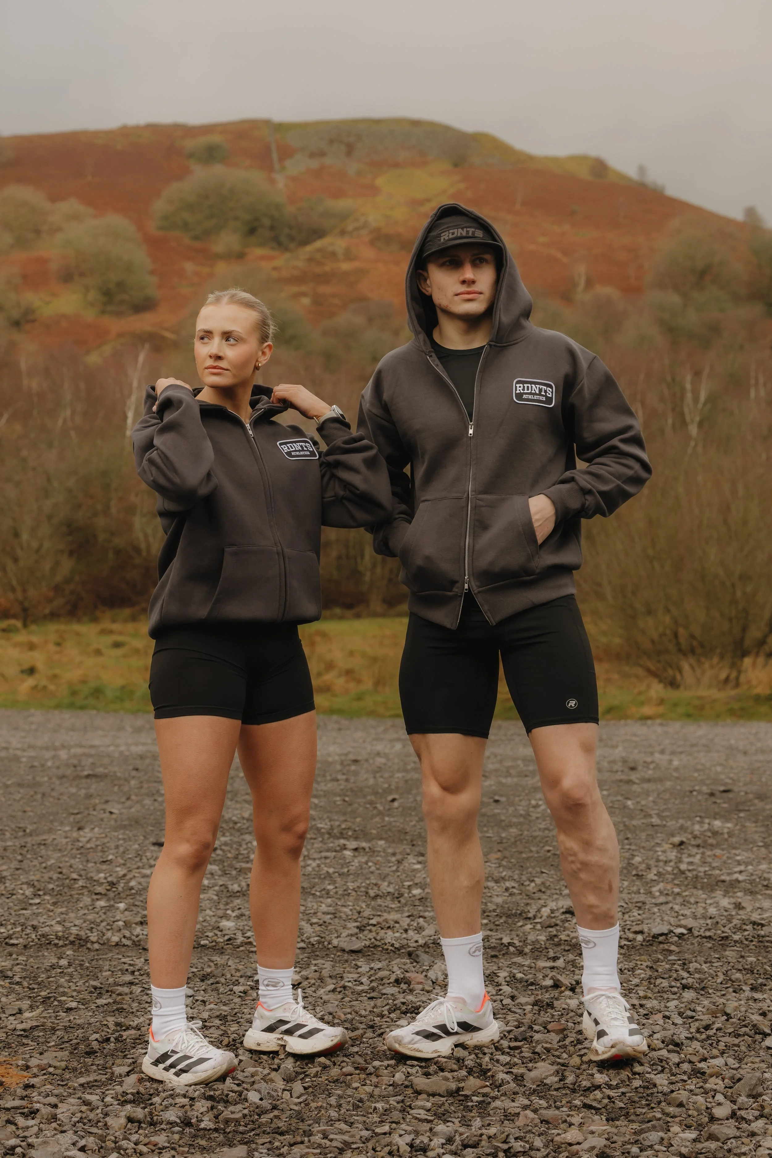 Two young adults posing outdoors on a rocky surface with hills and trees in fall colors in the background. They are wearing matching dark gray hoodie jackets and black athletic shorts.