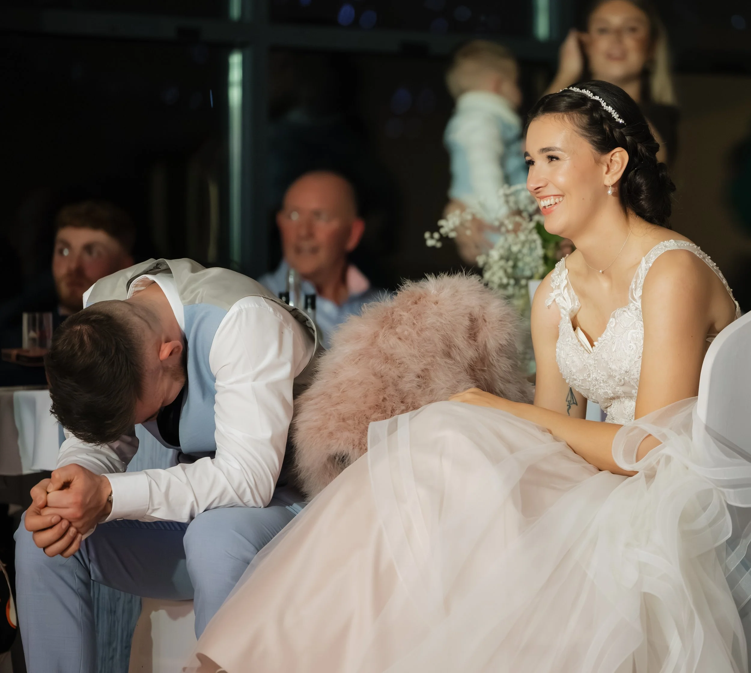 A bride in a white wedding dress sitting at a wedding reception, smiling, while a man, likely the groom, bows his head in front of her. In the background, guests are seated, watching the scene, and a woman holding a small child can be seen. The setti
