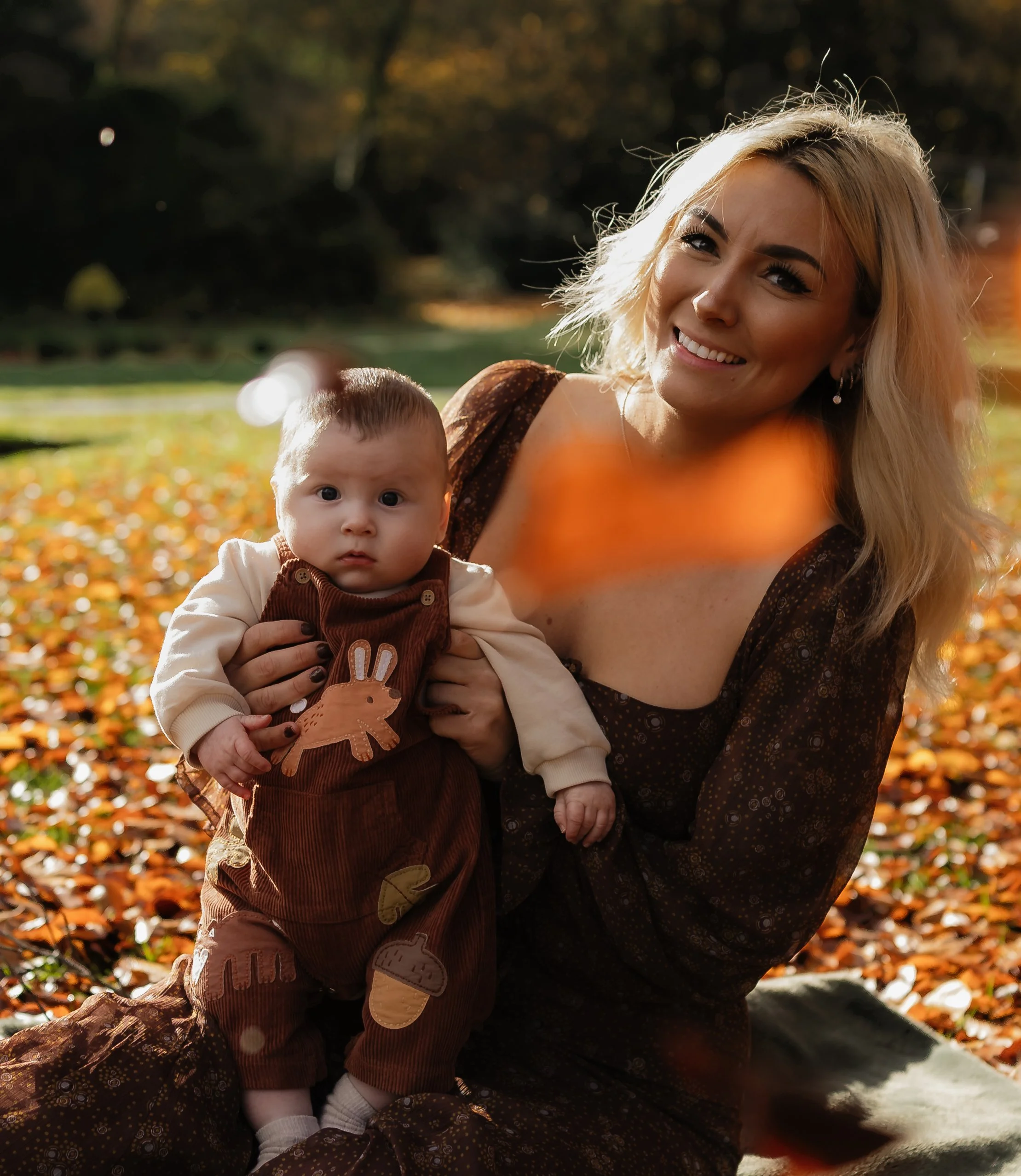 A woman with blonde hair and a baby outdoors in a park during autumn, with orange leaves on the ground and trees in the background.