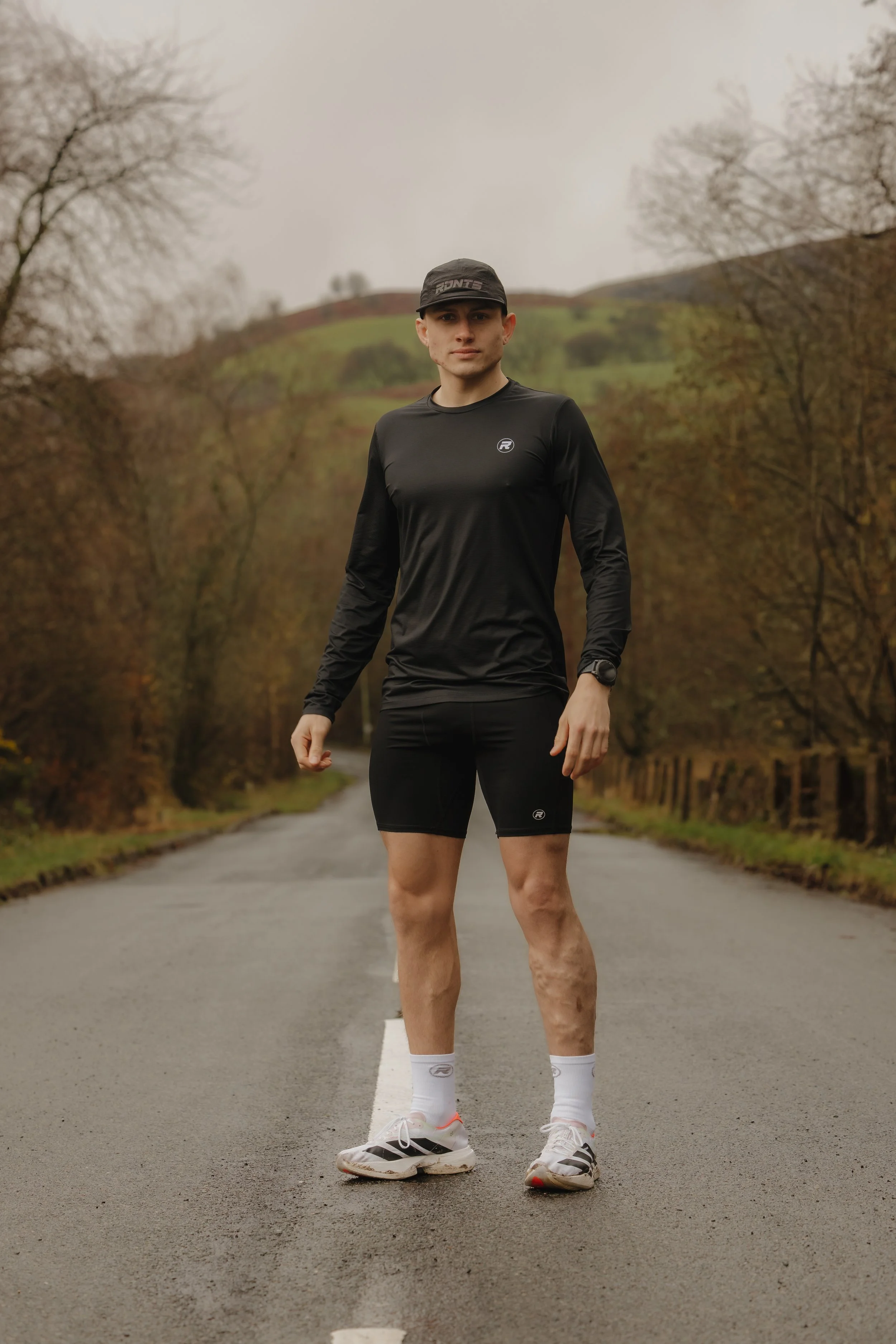 A young man standing on a rural road with autumn trees and hills in the background, wearing athletic clothing and sneakers.