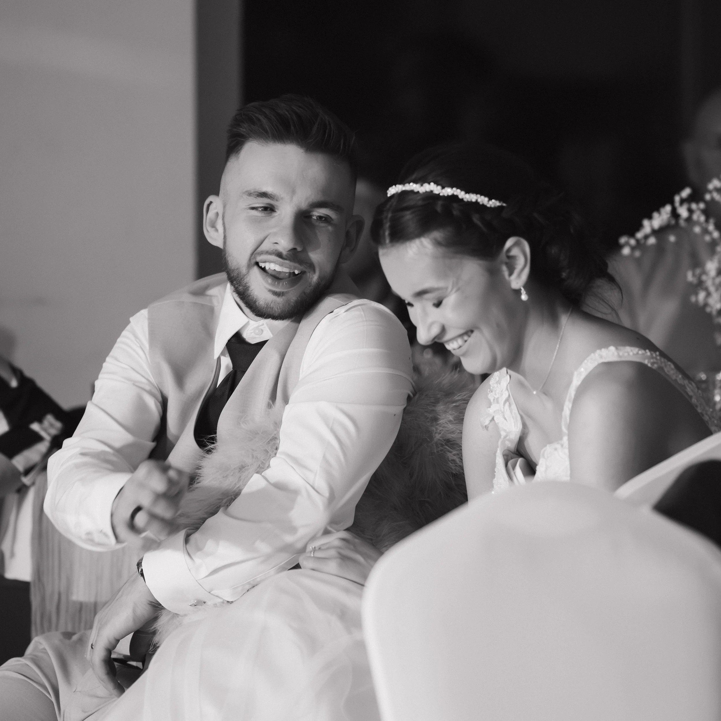 A black and white photo of a happy man and woman dressed in wedding attire, laughing together at a wedding reception.