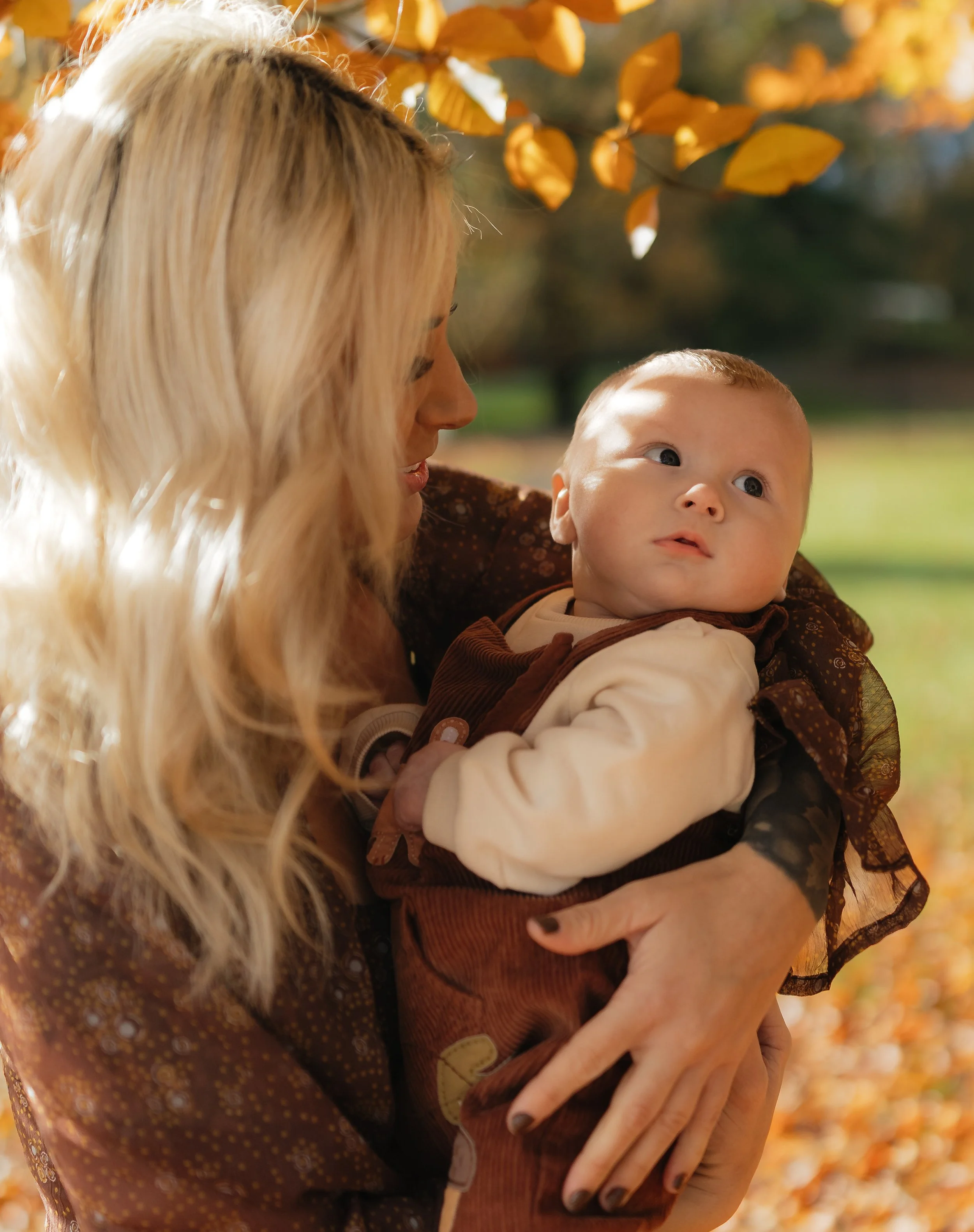 A woman with long blonde hair holding a baby in an outdoor setting with autumn leaves in the background.