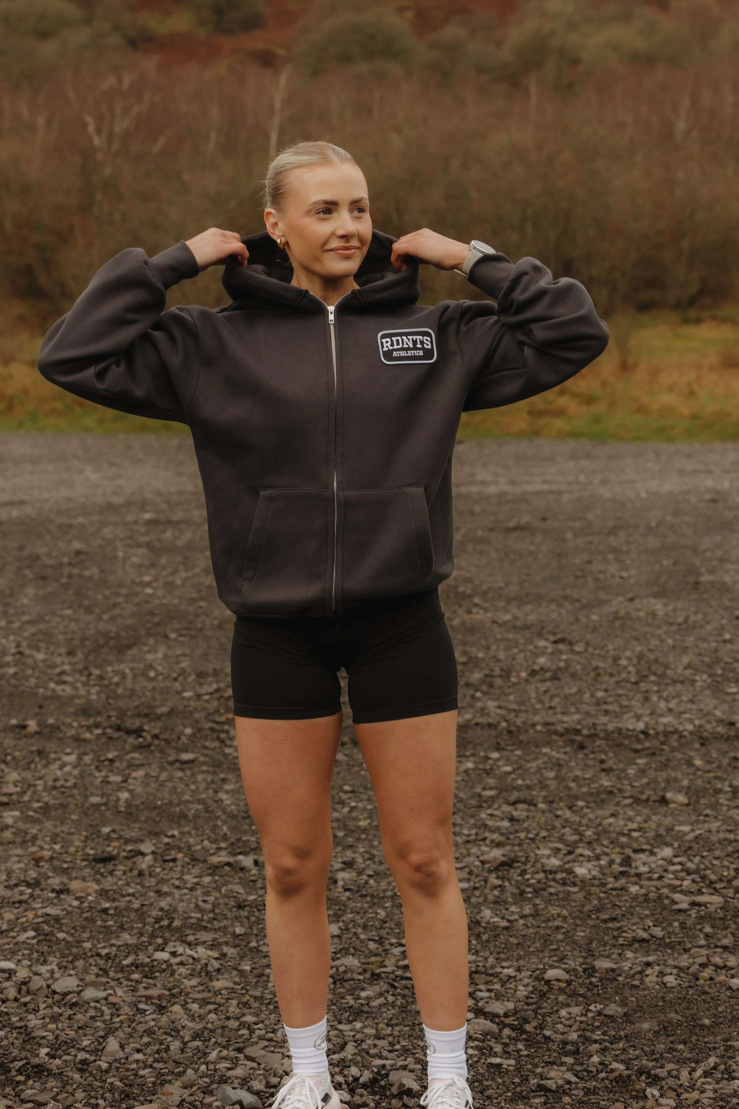 A woman in athletic clothing standing outdoors with a backdrop of trees and hills, adjusting her hoodie.