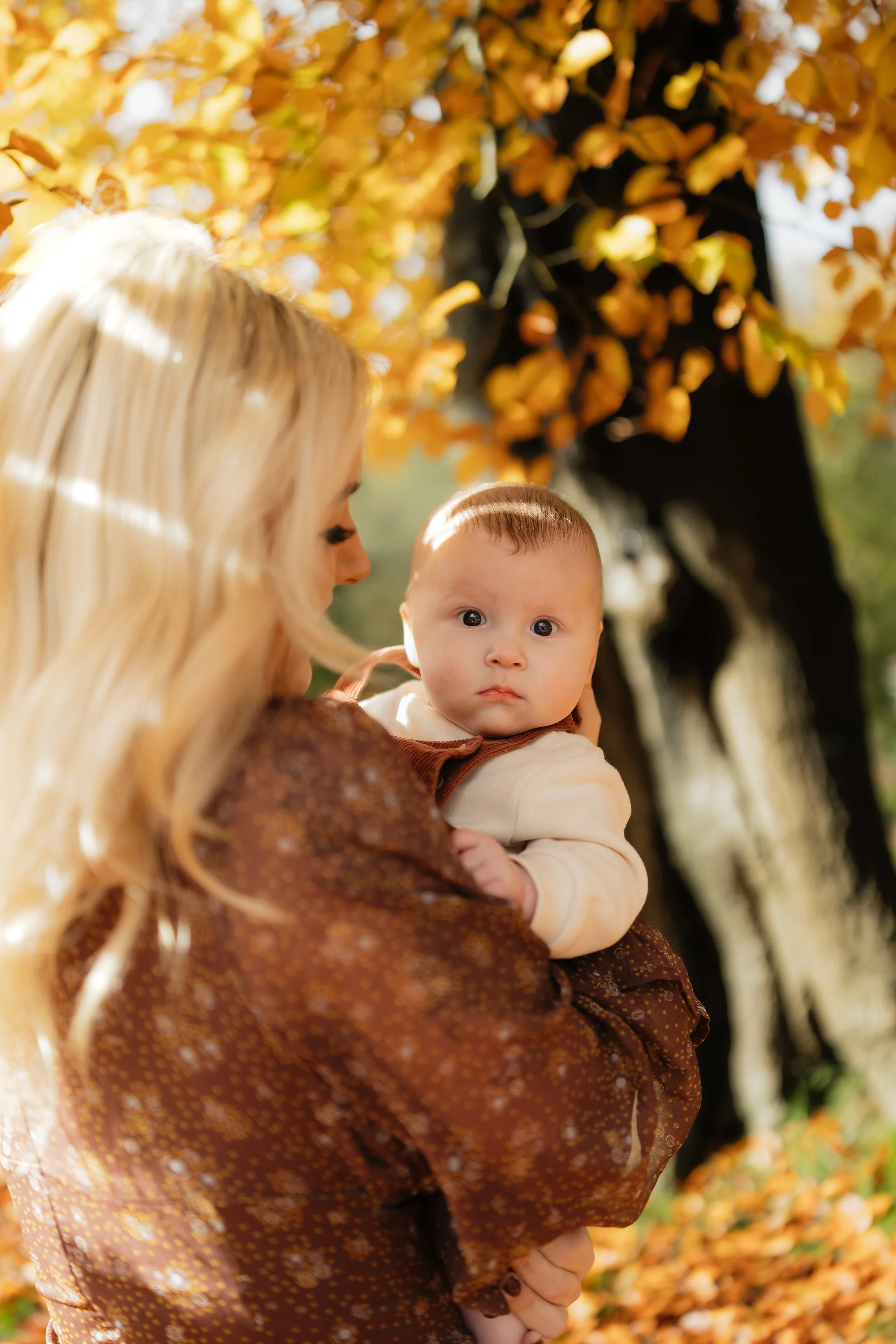 A woman holding a baby outdoors surrounded by fall leaves and trees.