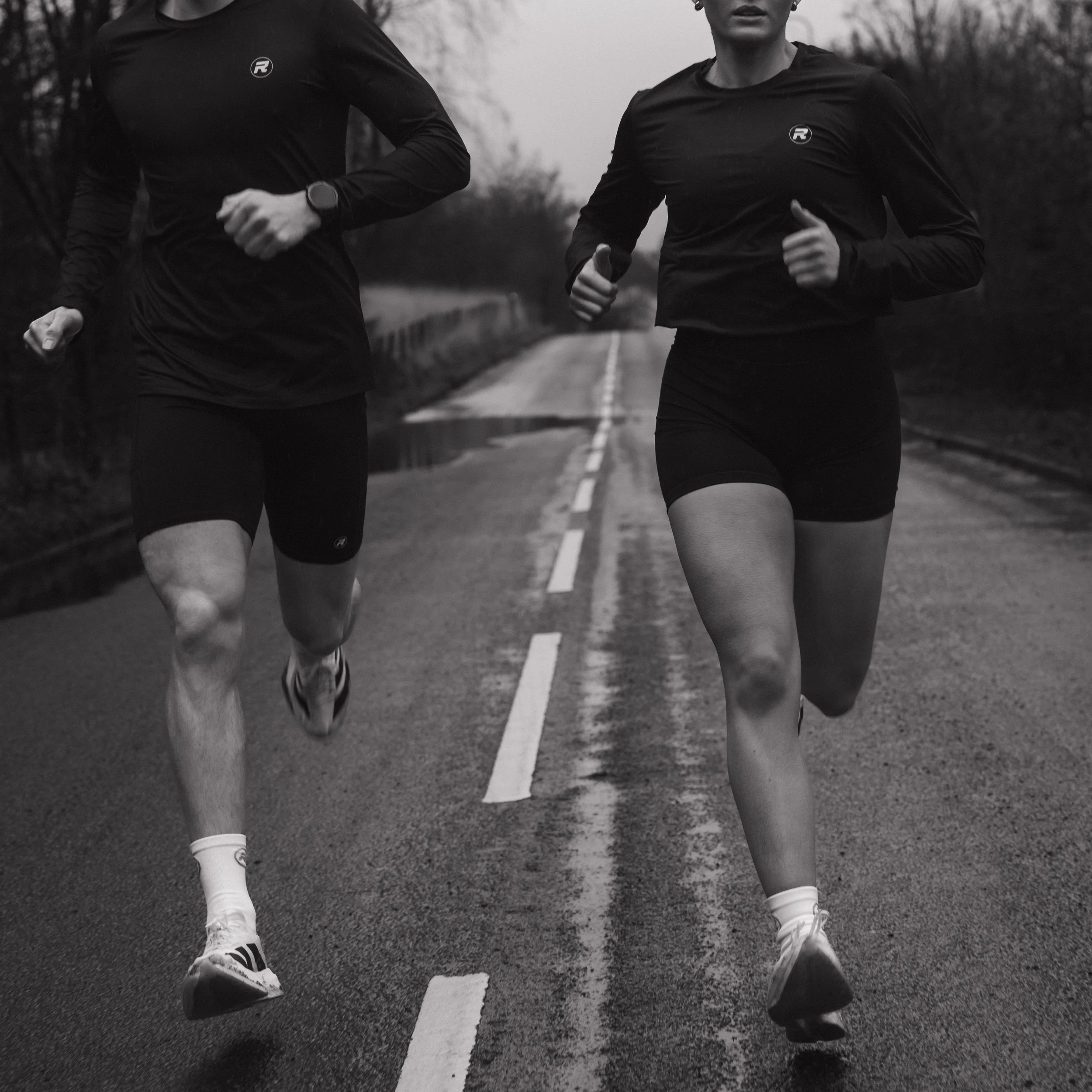 Black and white photo of a man and woman running on a rural road, mid-stride, wearing athletic clothing.