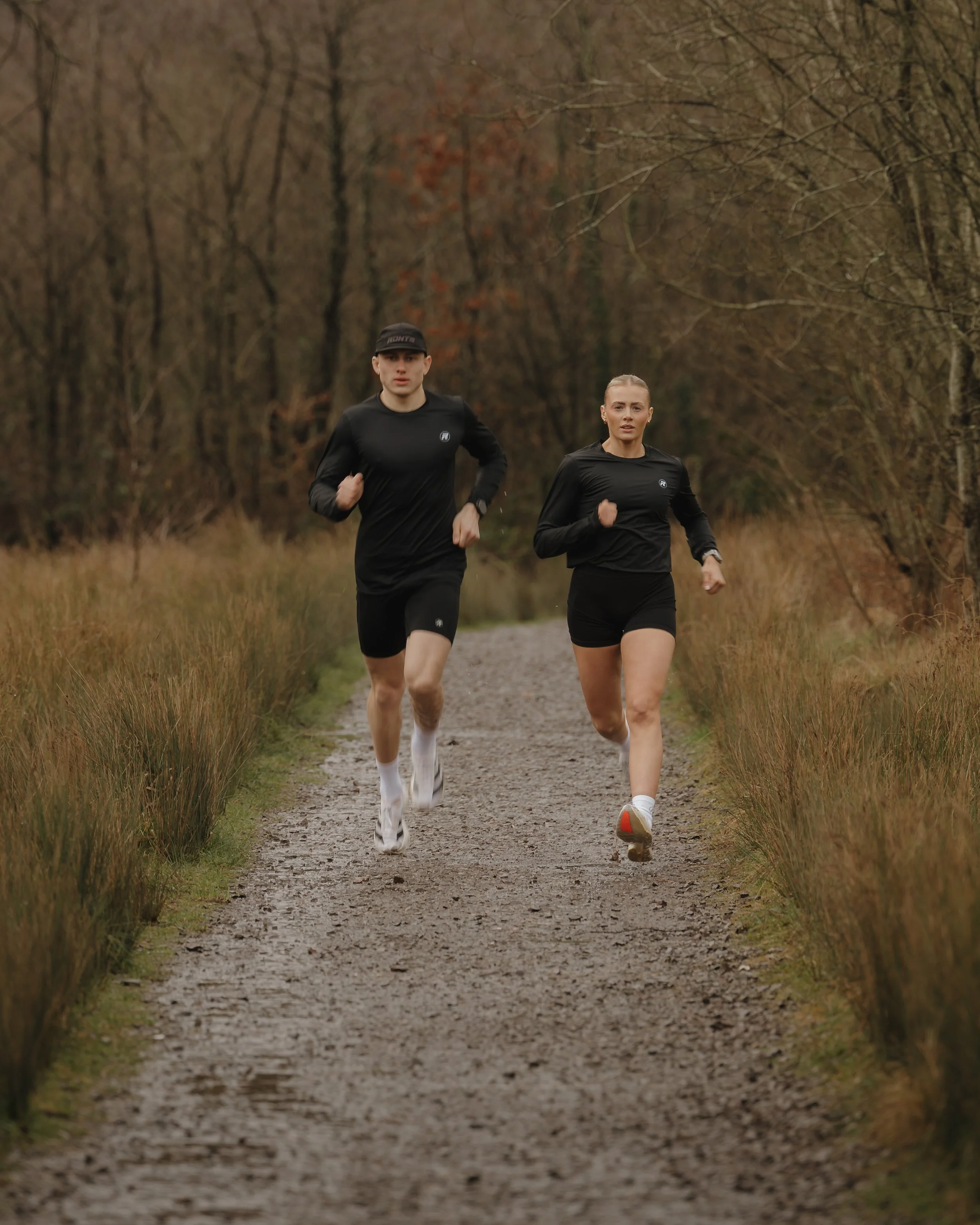 A man and woman running on a dirt trail through a wooded area with bare trees and grass on either side.