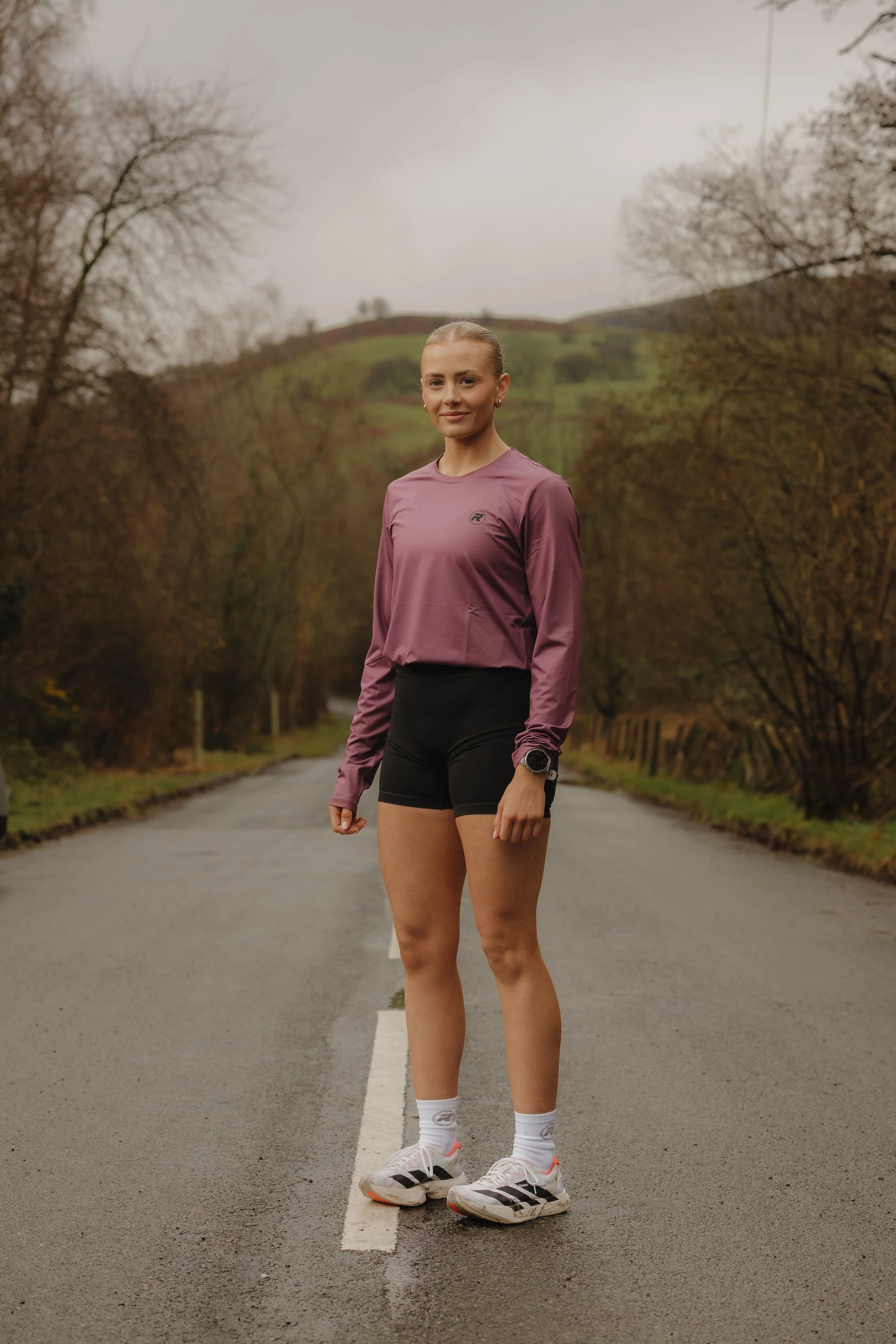 A young woman standing on a rural road surrounded by trees with a hill in the background, dressed in athletic wear including a pink long-sleeve shirt, black shorts, white running shoes, and white socks, with a watch on her left wrist.