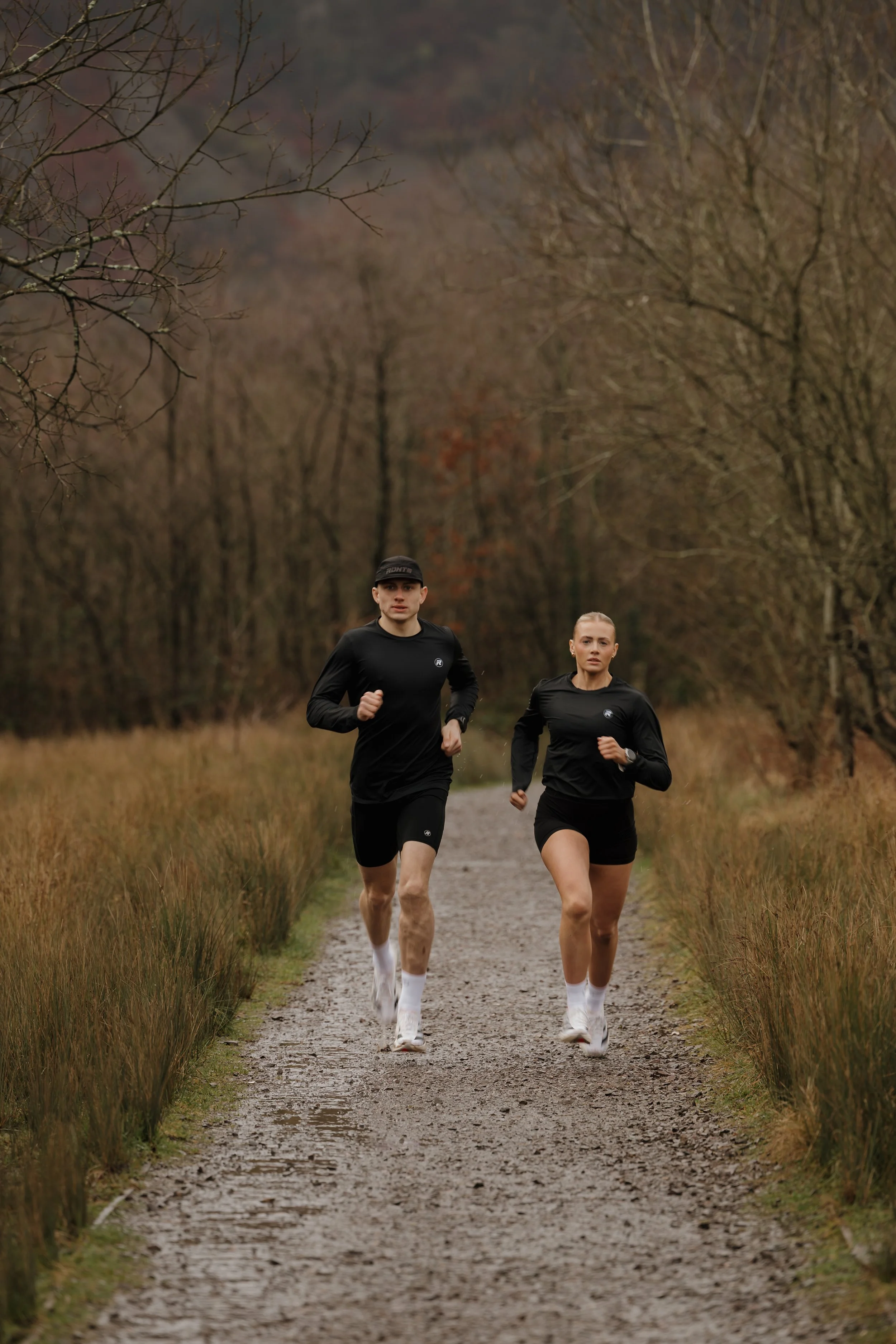 Two people running on a dirt trail through a forested area with trees and grass on either side.