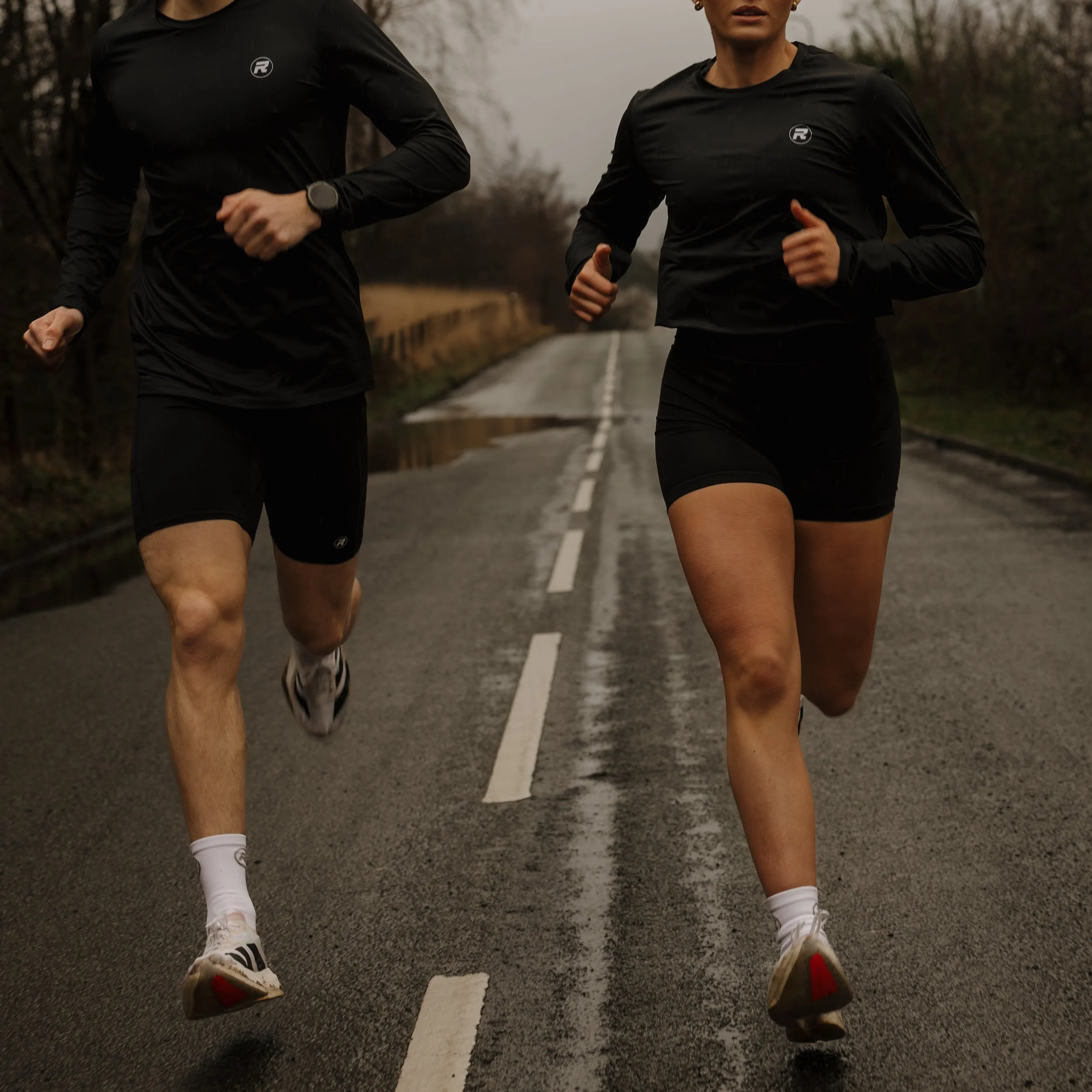 Two runners running on a wet road in an outdoor setting with trees and overcast sky.