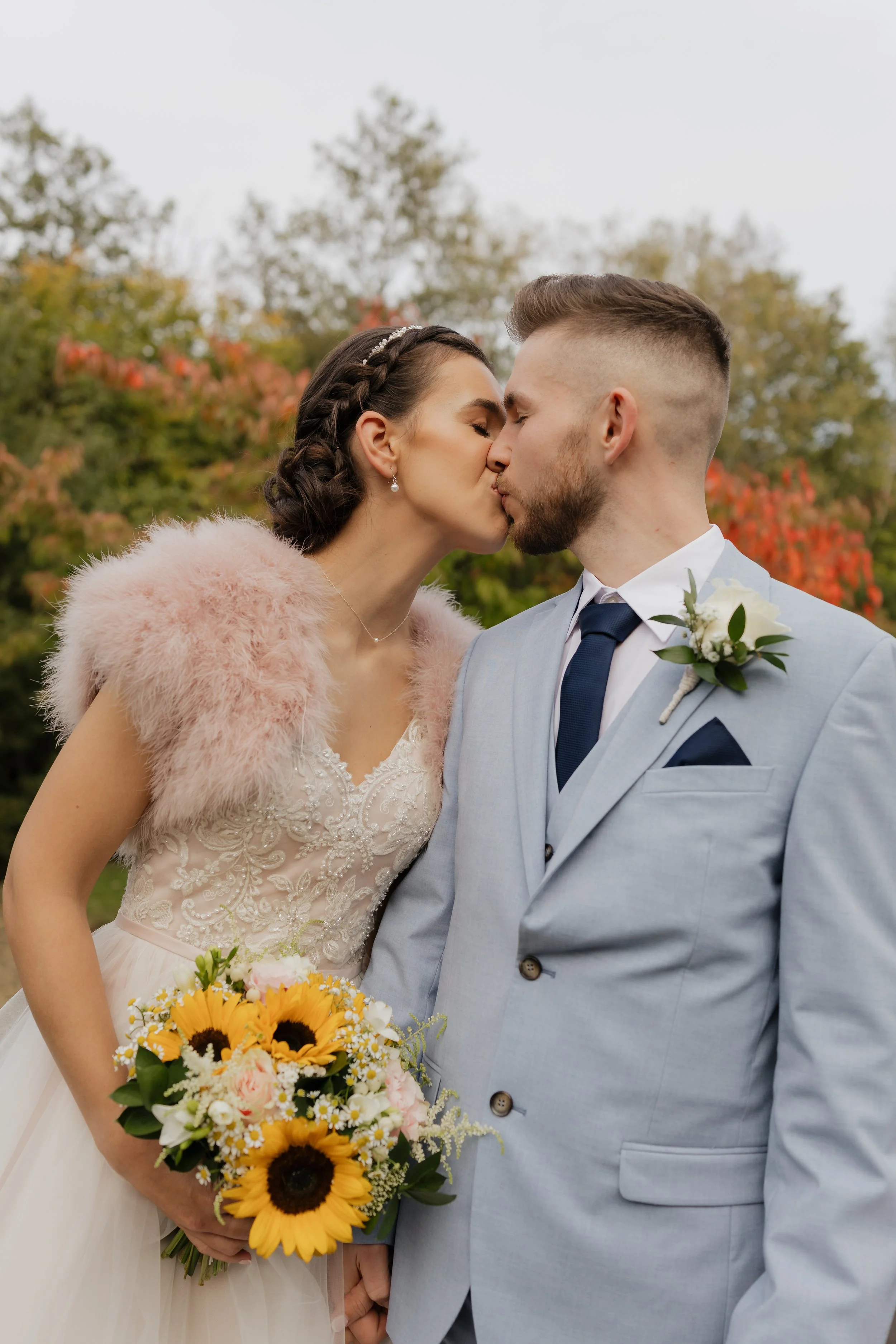 A bride and groom kissing outdoors on their wedding day, with autumn trees in the background. The bride is wearing a white lace dress with a pink faux fur shawl and holding a bouquet of sunflowers and other flowers. The groom is in a light gray suit 