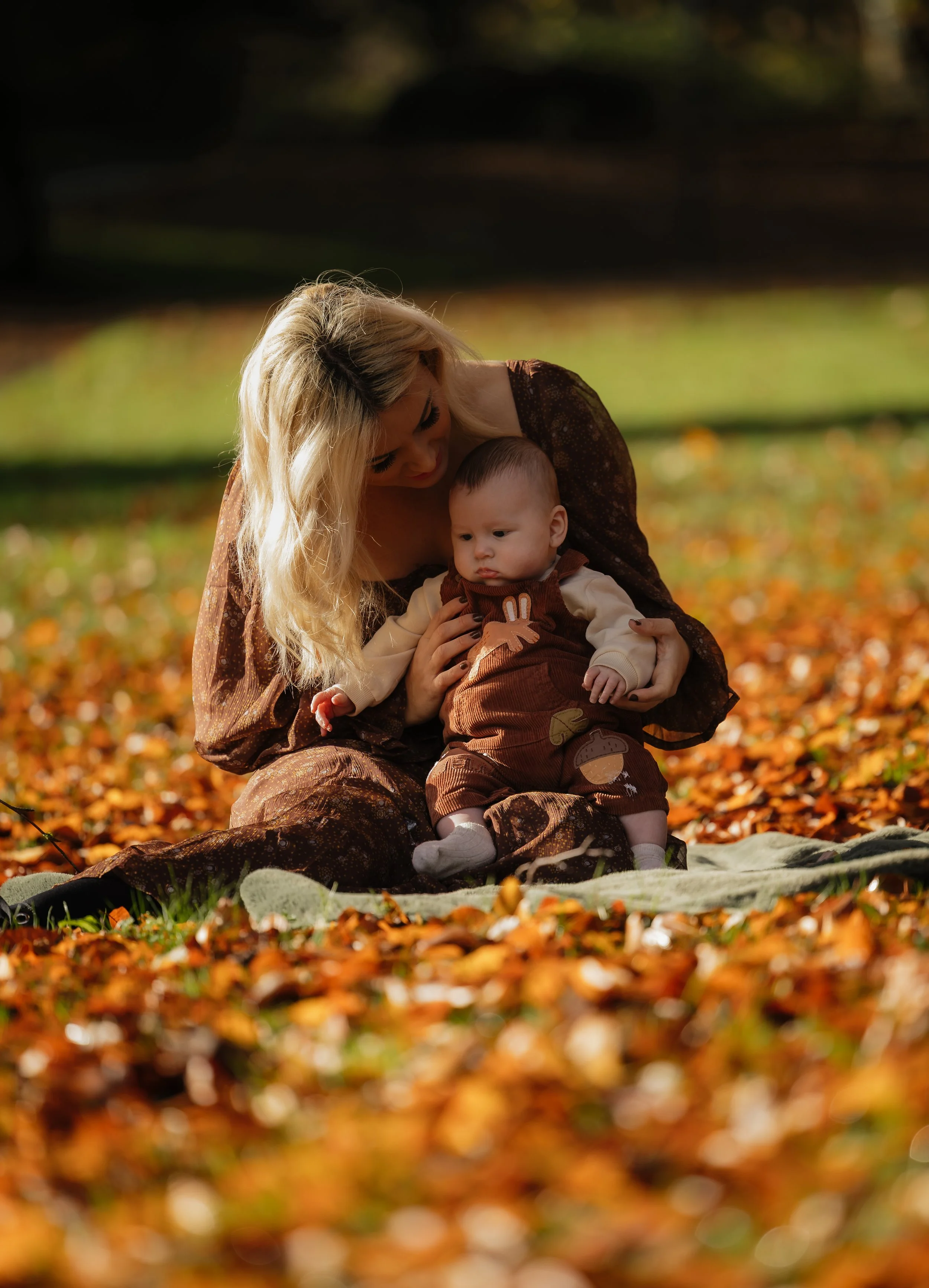 A woman with long blonde hair sitting on a blanket with a young child on her lap in an outdoor park during autumn, with fallen leaves around them.