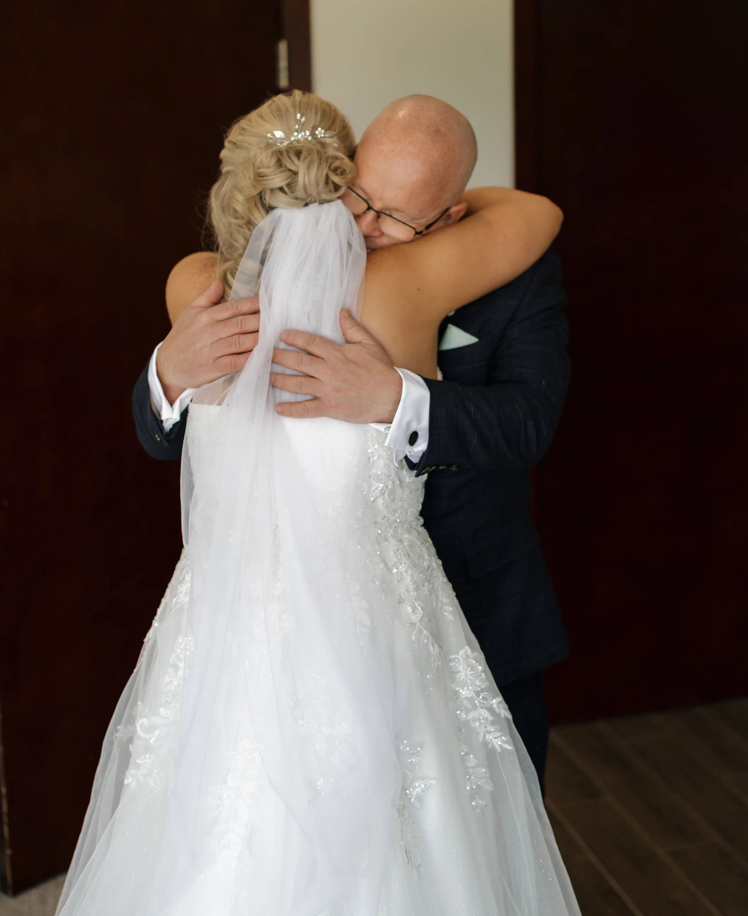 A bride and a man hugging each other, with the bride's back to the camera, wearing a white wedding dress and veil, and the man wearing a dark suit with a white shirt and glasses.