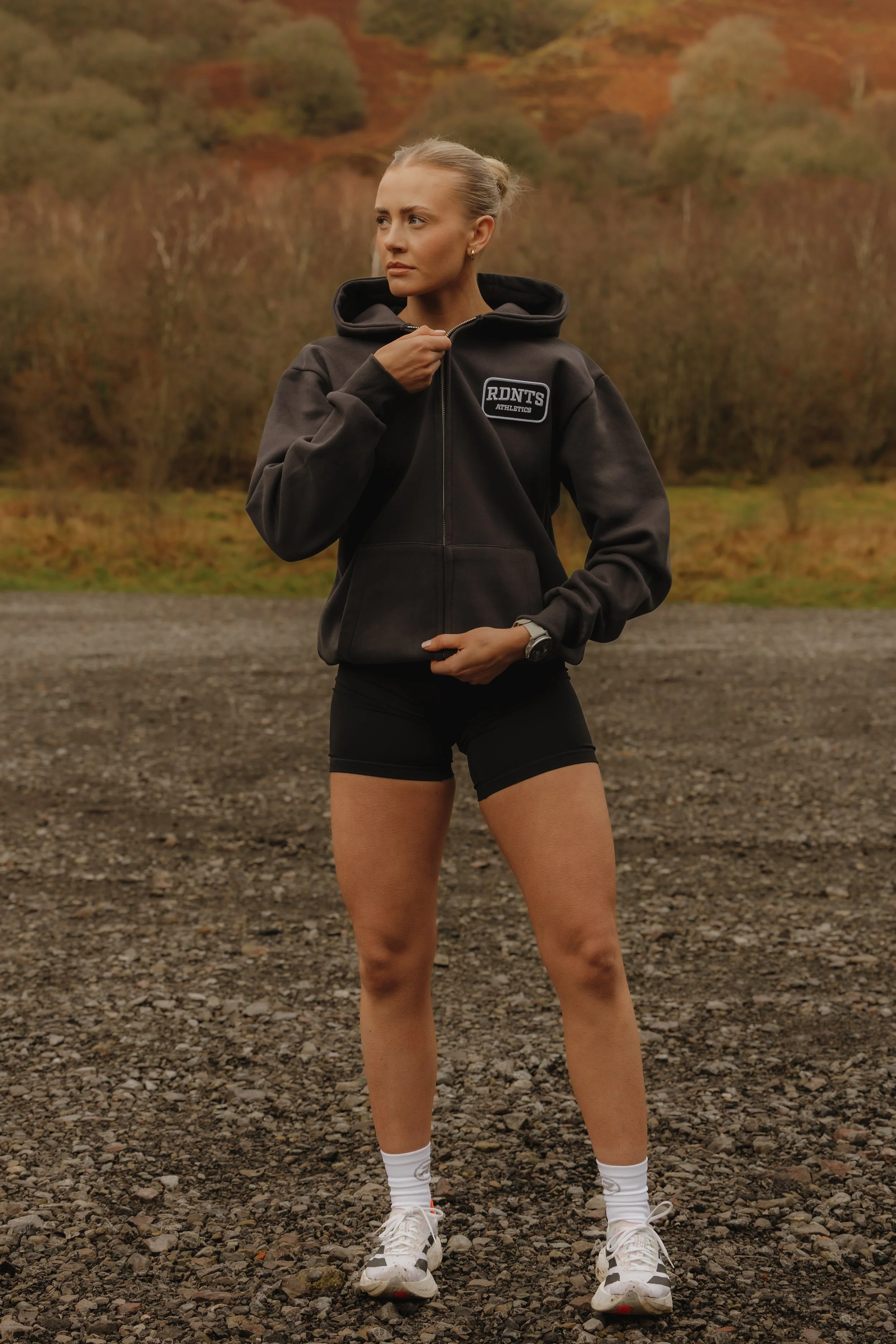 A woman in athletic gear standing outdoors on a gravel surface, with autumn trees and hills in the background, adjusting her black zip-up hoodie.