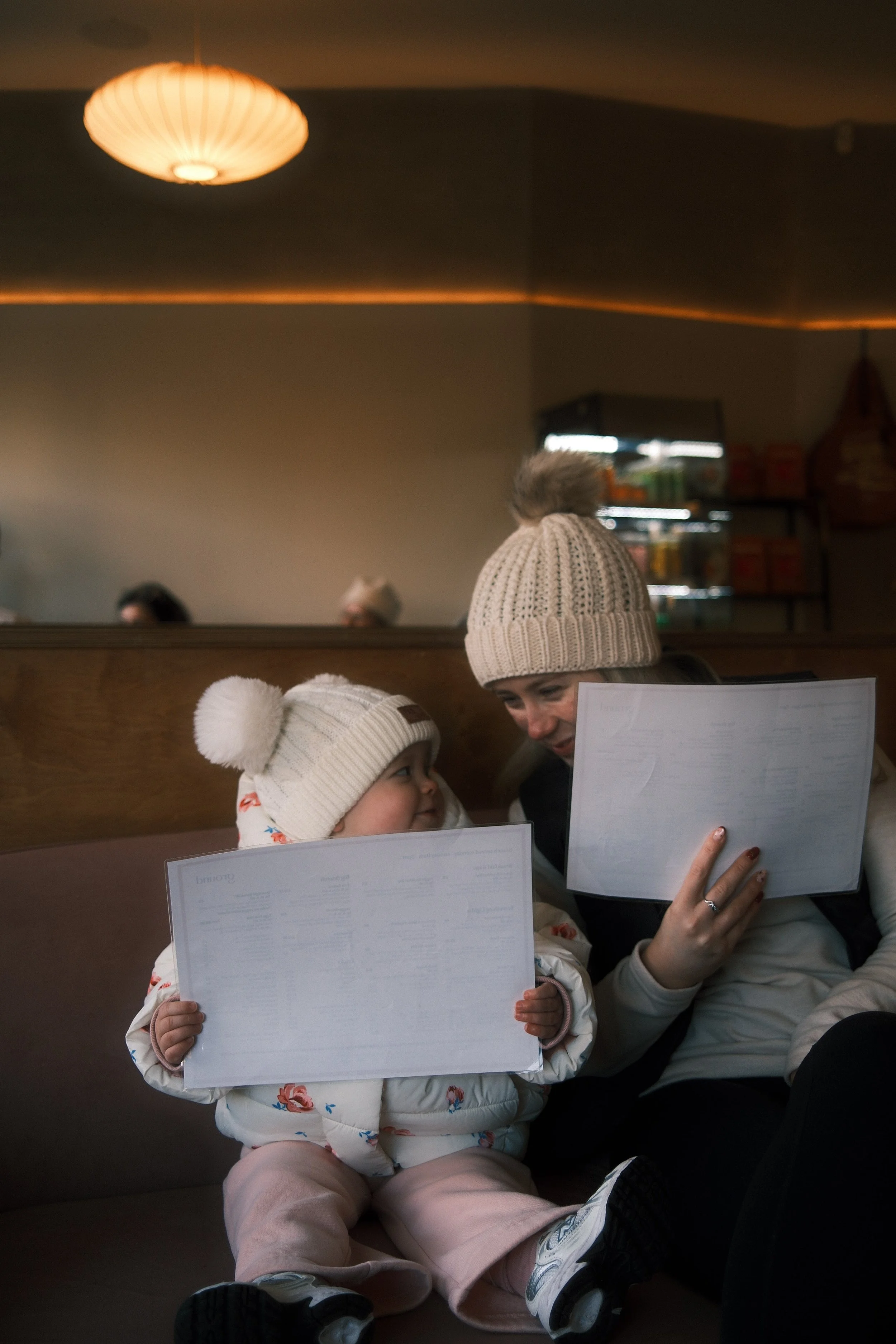 A woman and a young girl are sitting together in a restaurant, both holding menus. They are dressed warmly in winter hats with pom-poms and cozy clothing, smiling at each other.