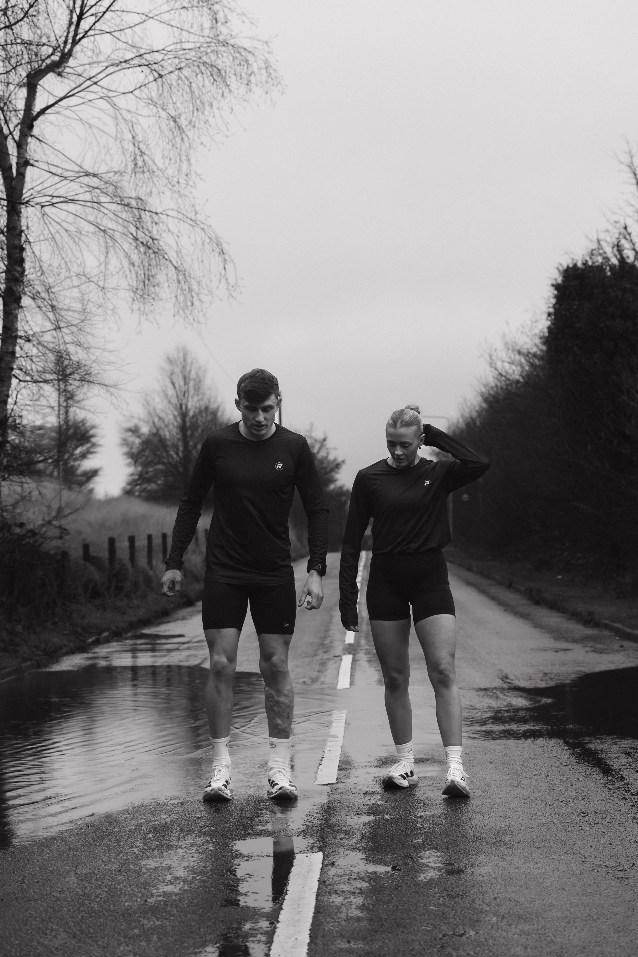 Two young people walking on a wet, empty road with puddles, surrounded by trees, in black and white.