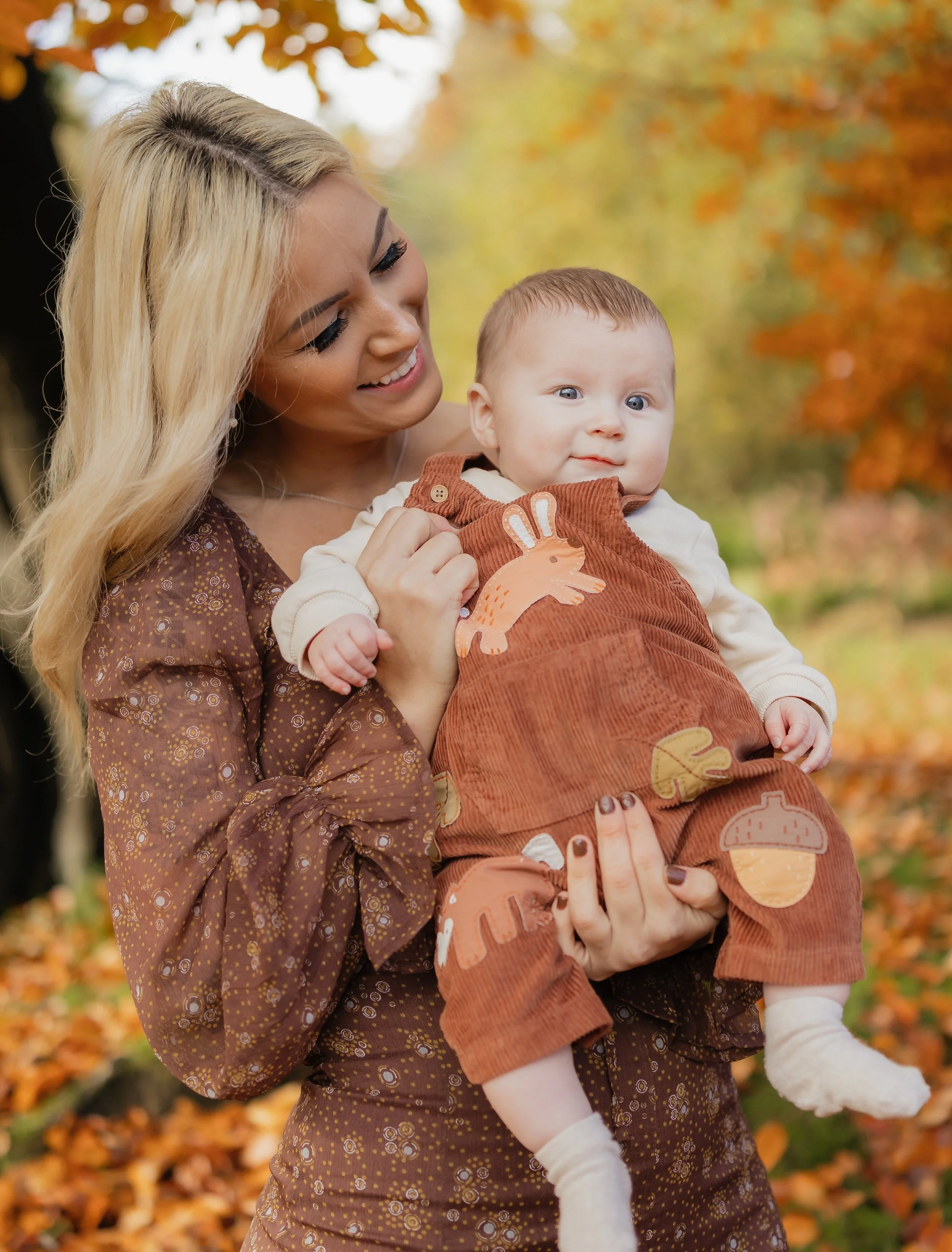 A woman holding a baby outdoors during fall with yellow and orange leaves in the background.