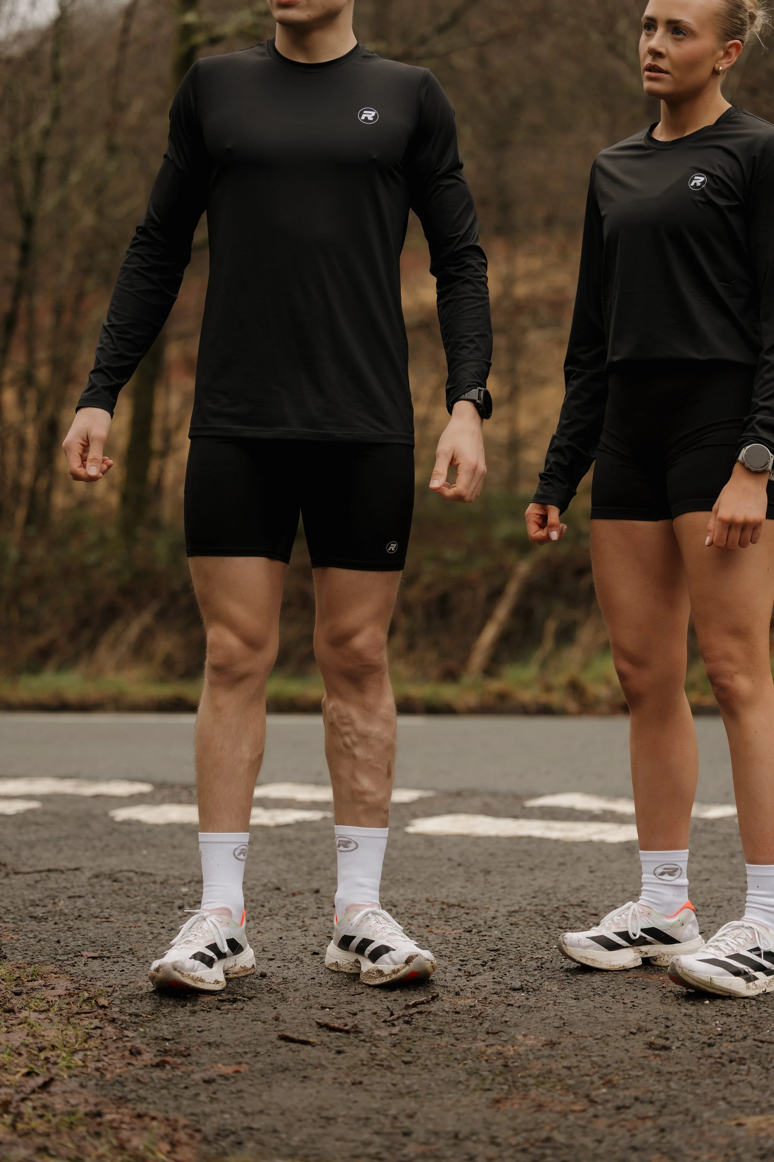 Two runners standing outdoors on a road, dressed in black athletic gear, wearing white running shoes and socks, with a forest background.