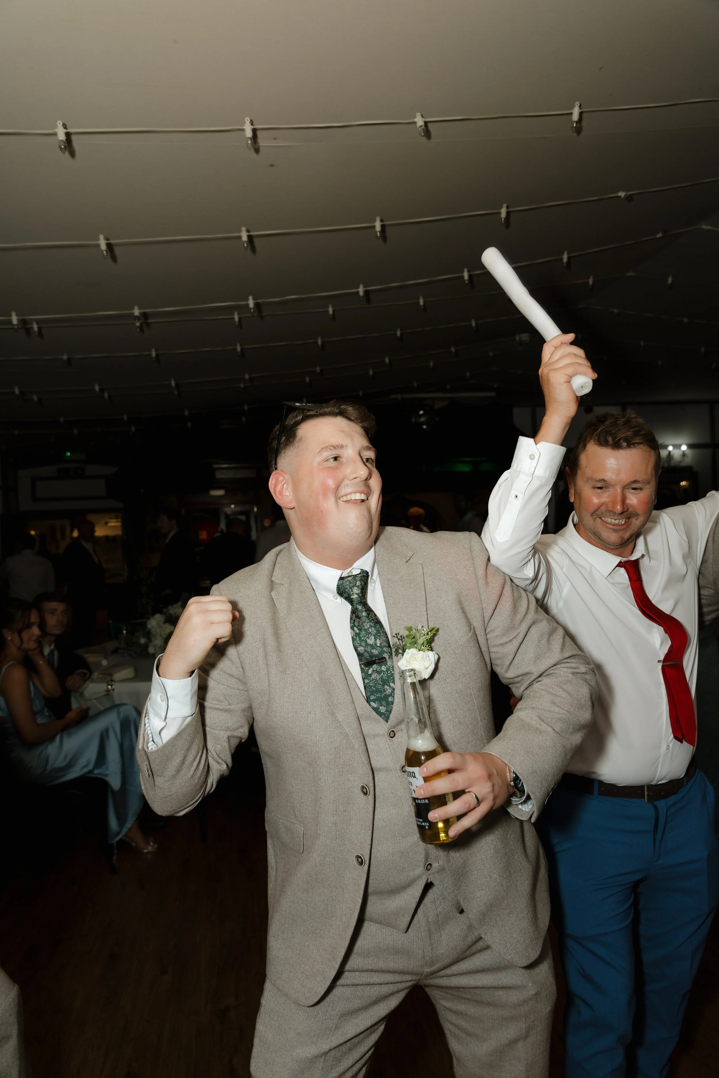 People celebrating at a party or wedding reception, with two men in the foreground cheering, one holding a beer and the other a foam stick, smiling and dressed in formal attire.