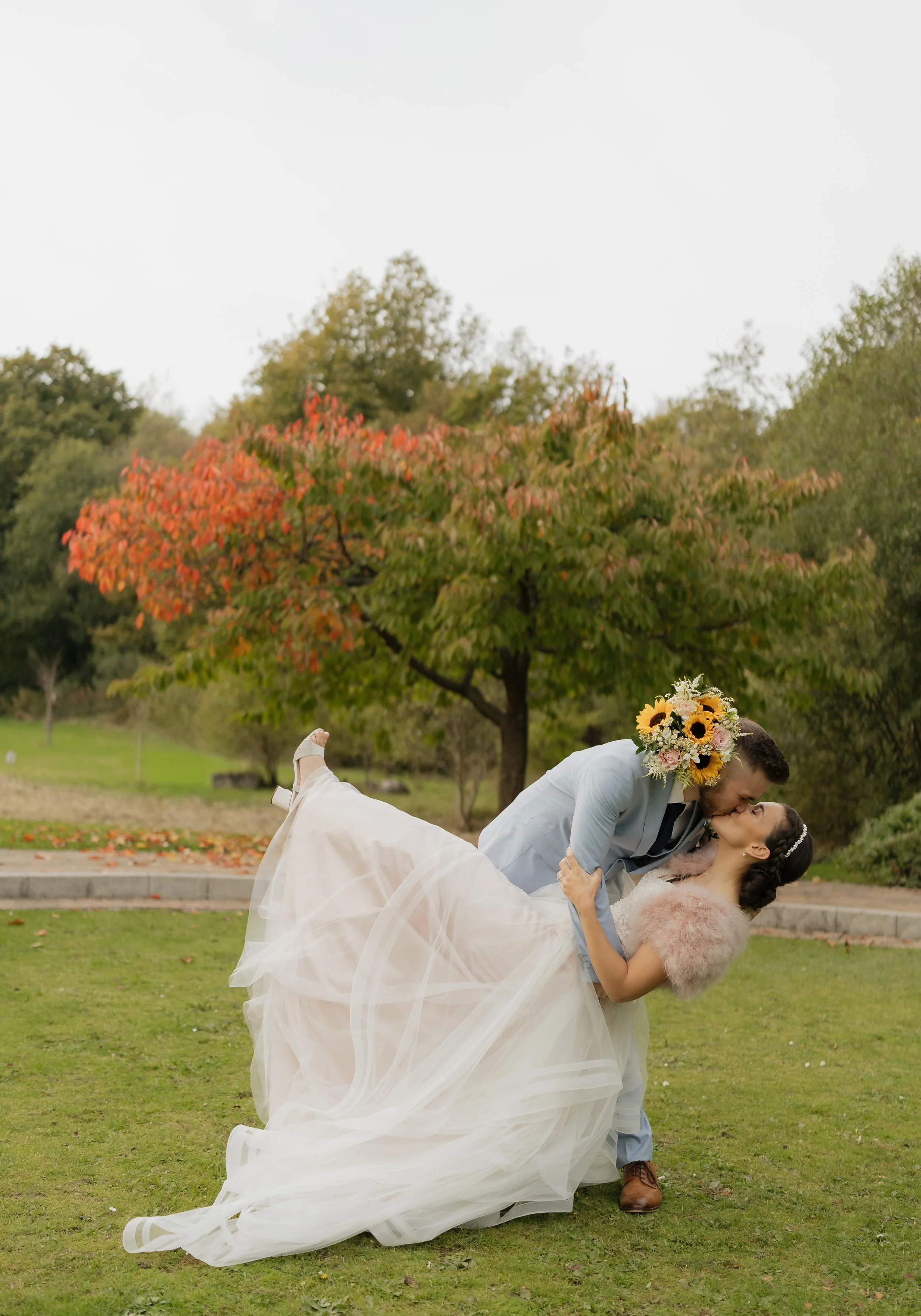 A bride and groom sharing a kiss outdoors on a grass field, with the groom holding the bride in a dip. The bride is wearing a white wedding dress with a tulle skirt and a pink fur shawl, and the groom is dressed in a light blue suit with brown shoes.