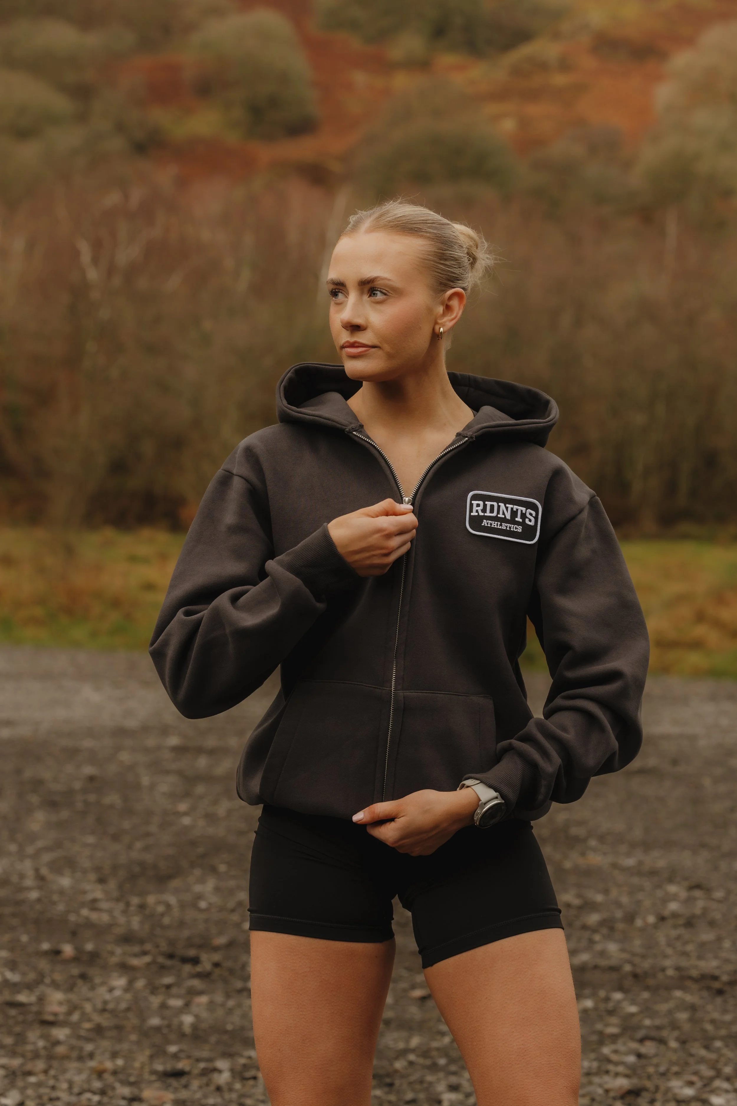A woman in a dark athletic hoodie and black shorts outdoors, adjusting her zipper with a wooded landscape in the background.