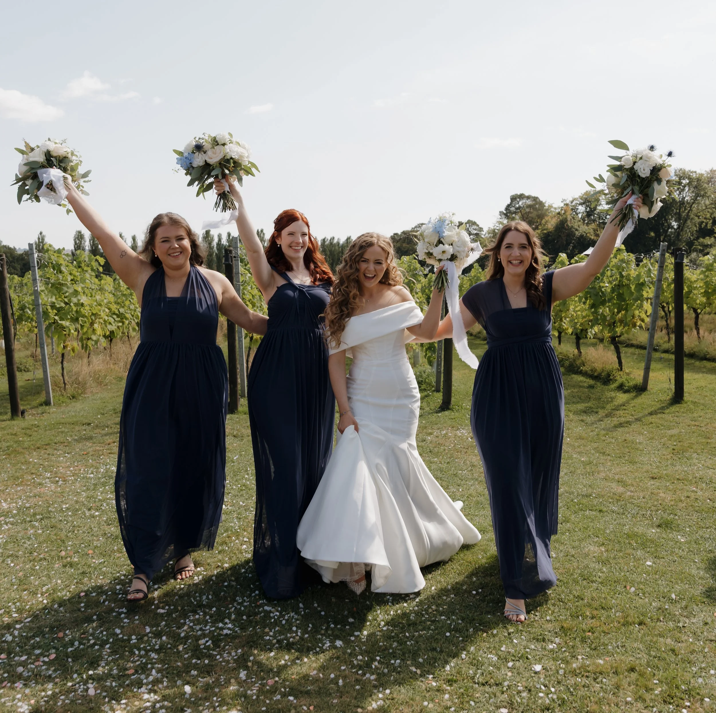 A bride in a white wedding dress walking outdoors with three women in navy blue bridesmaid dresses, holding bouquets, celebrating on a sunny day in a vineyard.
