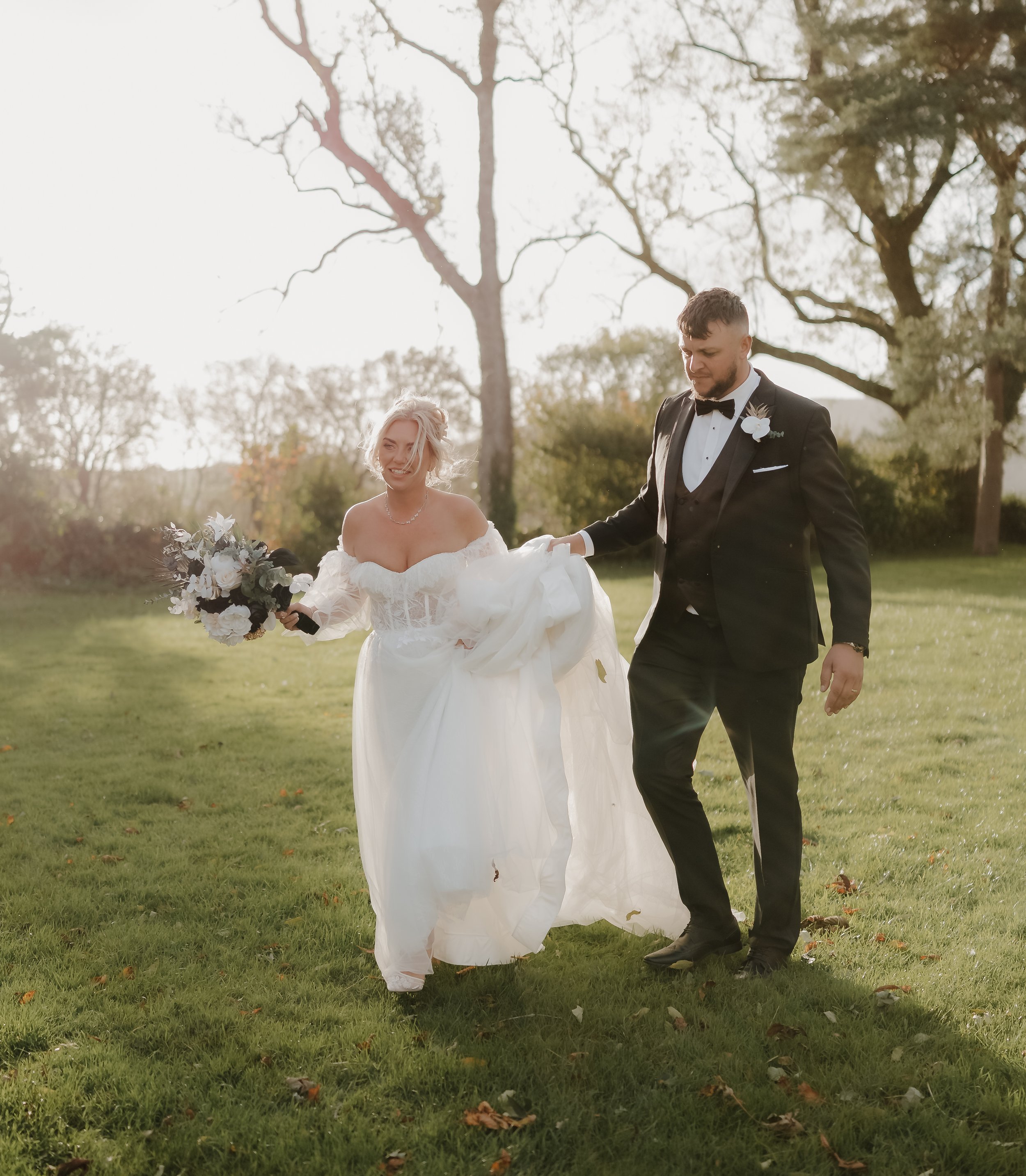 A bride and groom walking on a grassy field during their wedding, with the bride holding a bouquet of flowers and the groom lifting part of her dress, both smiling and dressed in wedding attire, under a backdrop of trees and sunlight.