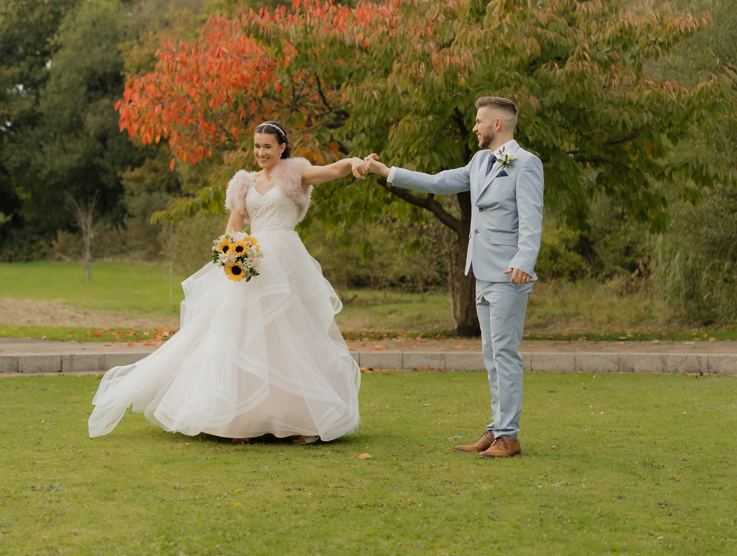 A bride and groom holding hands in an outdoor park with green grass and trees with autumn-colored leaves, smiling and happy.