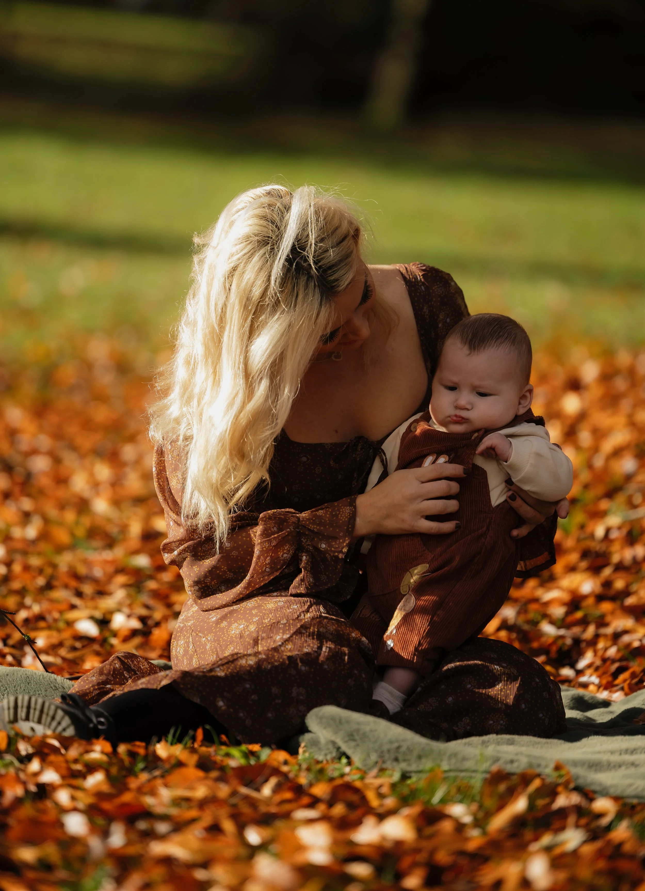 A woman with long blonde hair sitting on a blanket on autumn leaves, holding a baby in her arms, outdoors in a park.