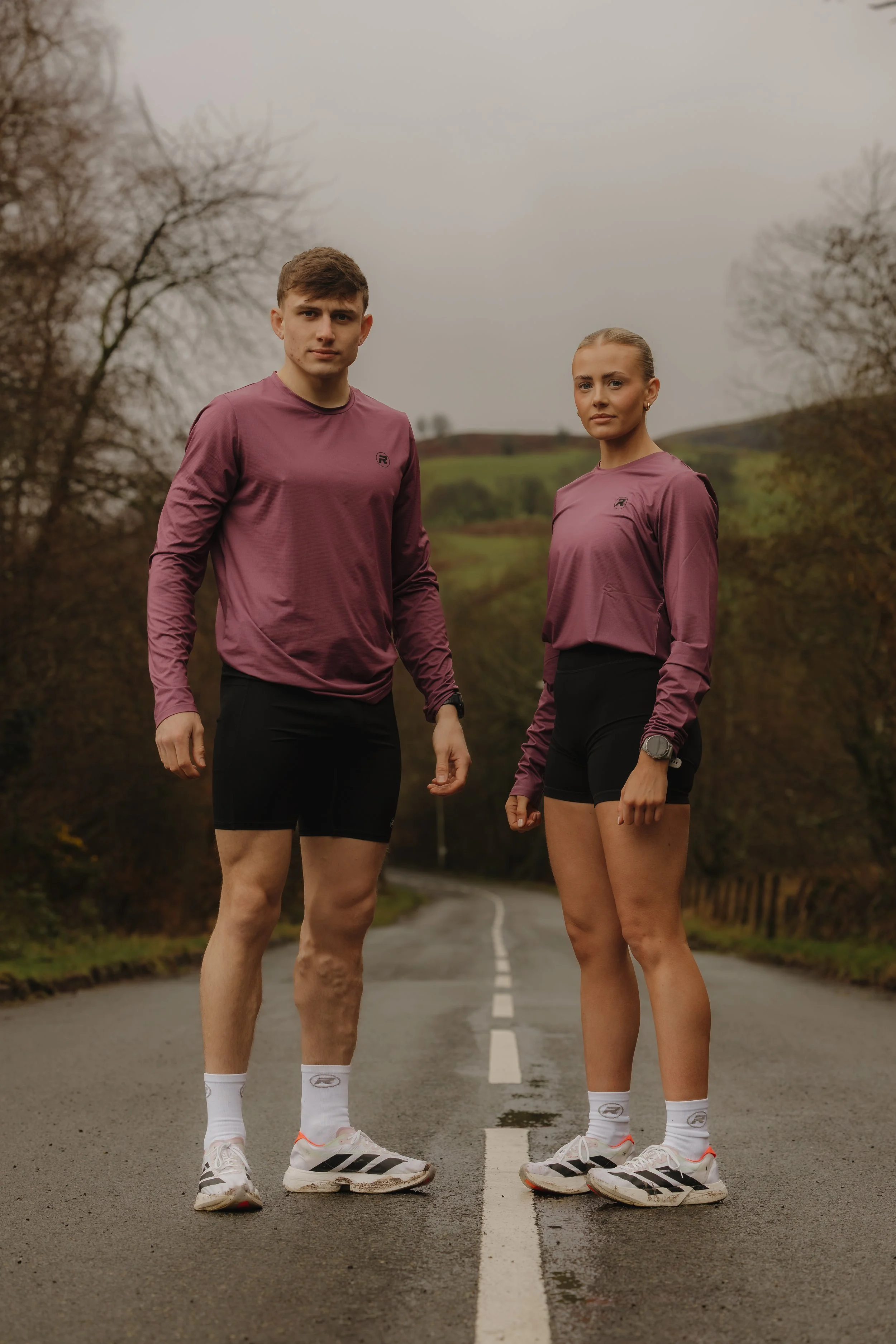 A young man and woman standing side by side on a deserted road in a rural area, dressed in matching athletic wear, surrounded by trees and hilly landscape under an overcast sky.