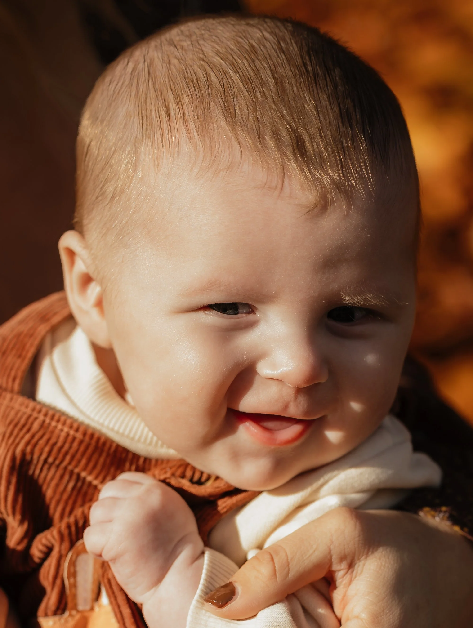 Close-up of a smiling baby with short hair, lying outdoors in autumn leaves, wearing a brown vest over a cream-colored shirt.