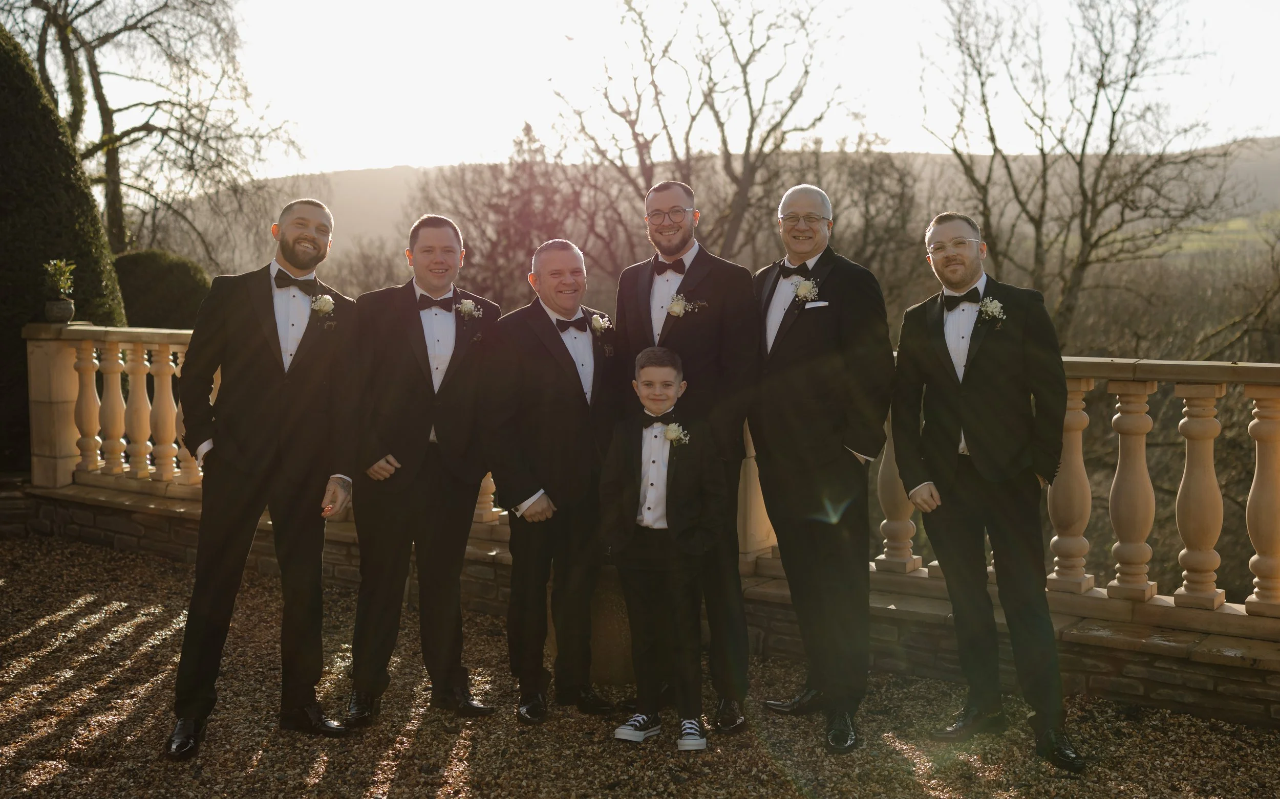 Group of men and a boy in tuxedos posing outdoors at sunset, trees in the background, on a terrace with a balustrade.