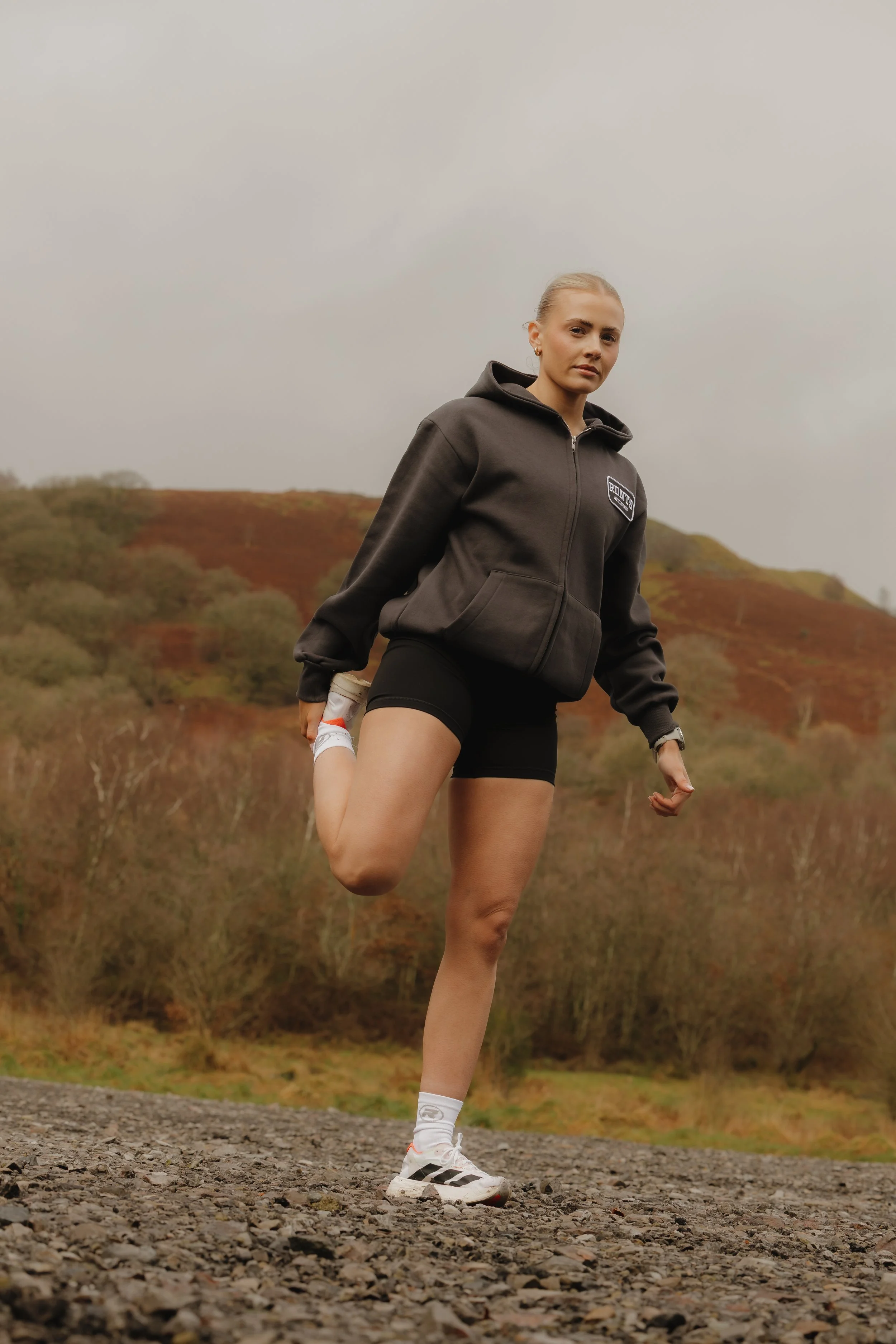 Woman in athletic wear stretching outdoors on rocky ground with hillside and trees in the background.