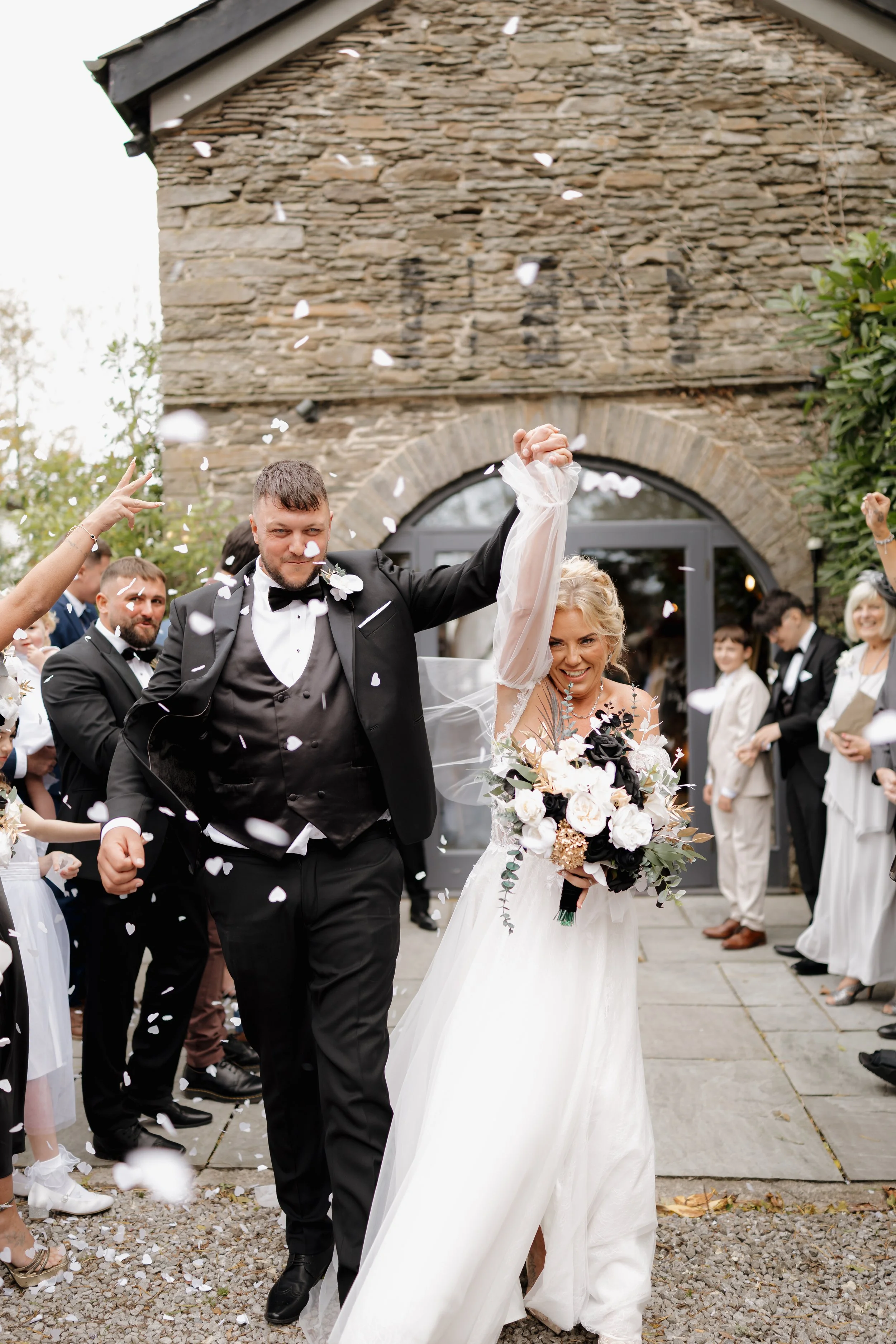 A bride and groom celebrating their wedding, holding hands and smiling as friends and family throw confetti during outdoor ceremony in front of a stone building.