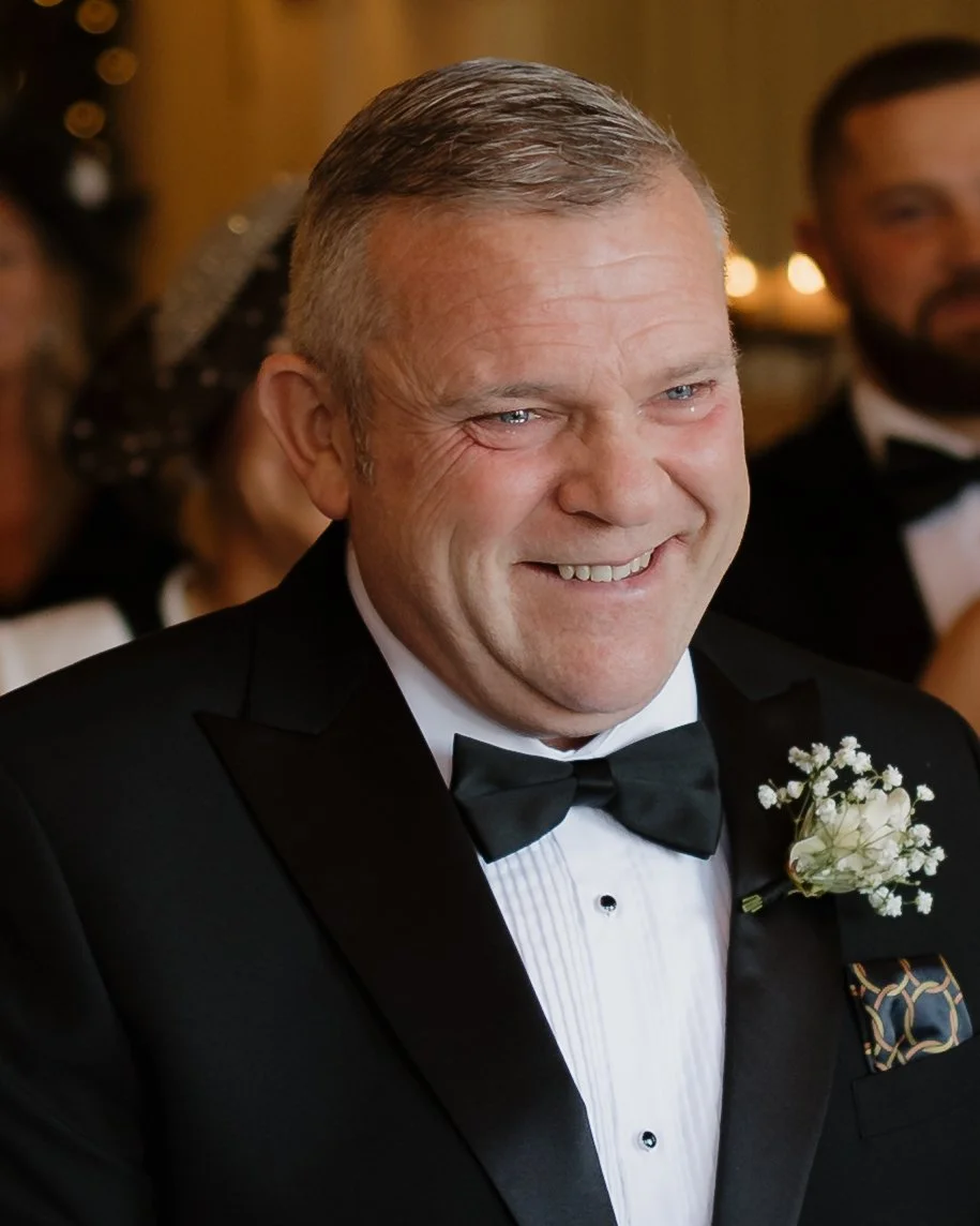 A smiling man in a tuxedo with a white boutonniere, attending a formal event or wedding, with other guests in the background.
