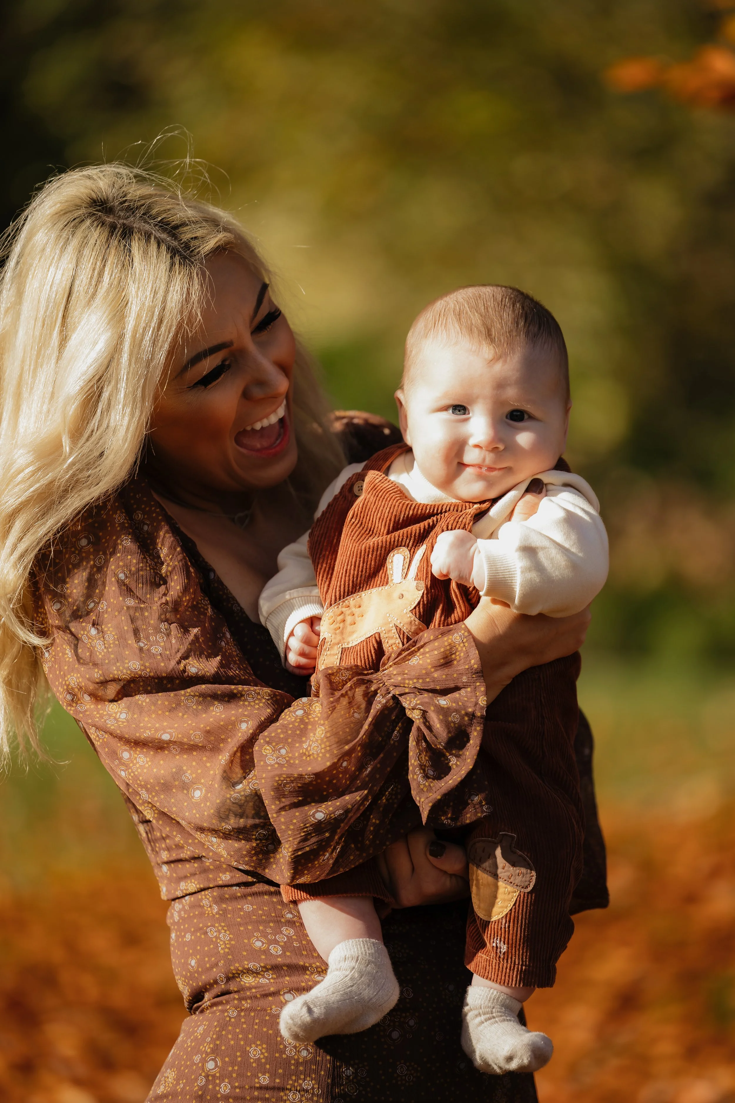 A woman with long blonde hair smiling and holding a baby outdoors in an autumn setting with fallen leaves.