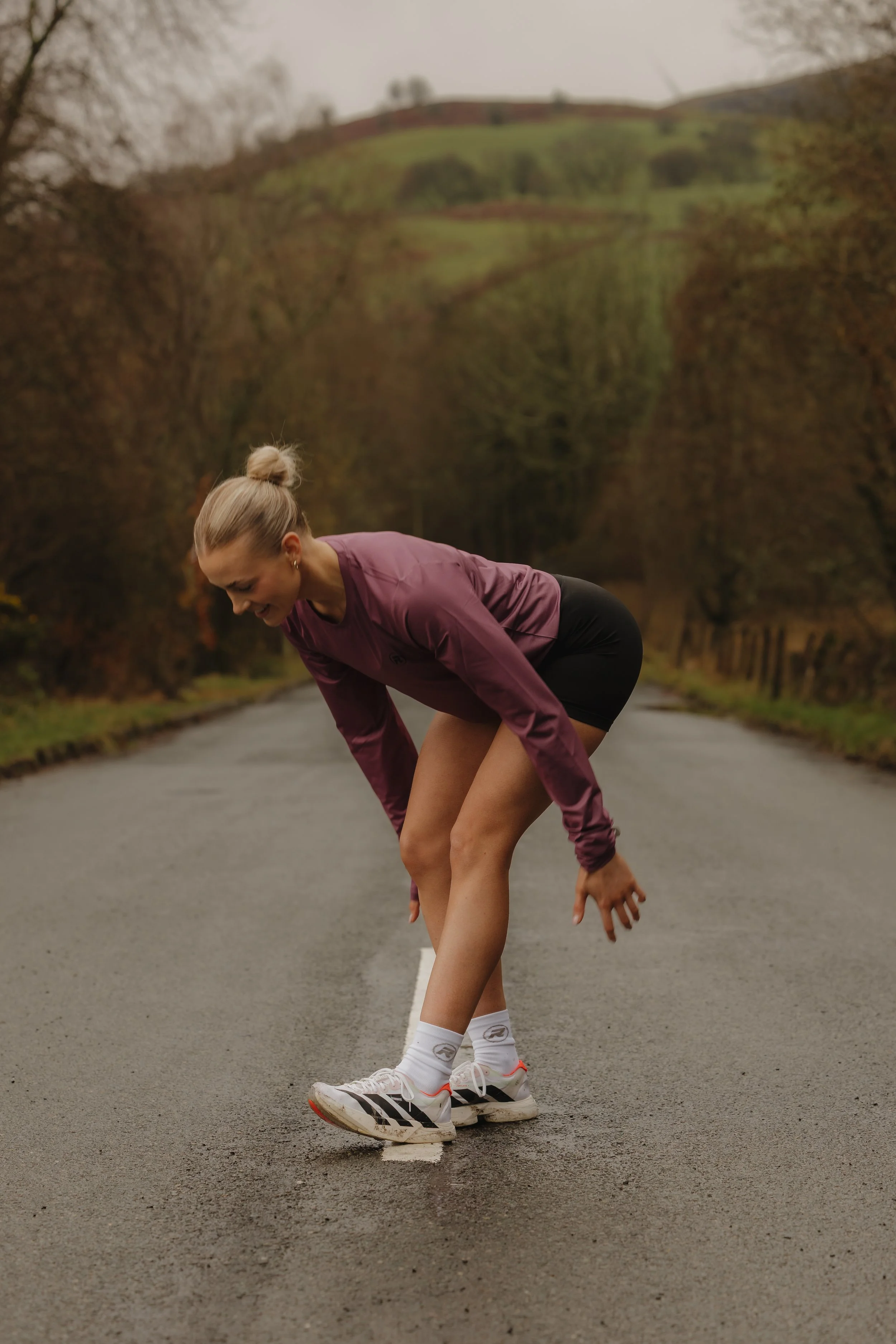 Woman in athletic clothing bending over on a road, with a background of trees and hills, clutching her ankle in pain.