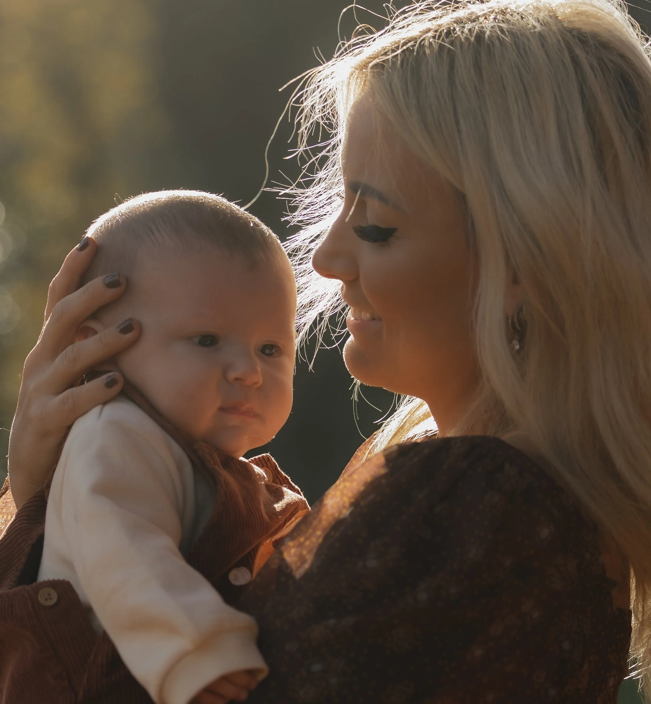 A woman with long blonde hair holding a baby close, both outdoors with sunlight in the background.