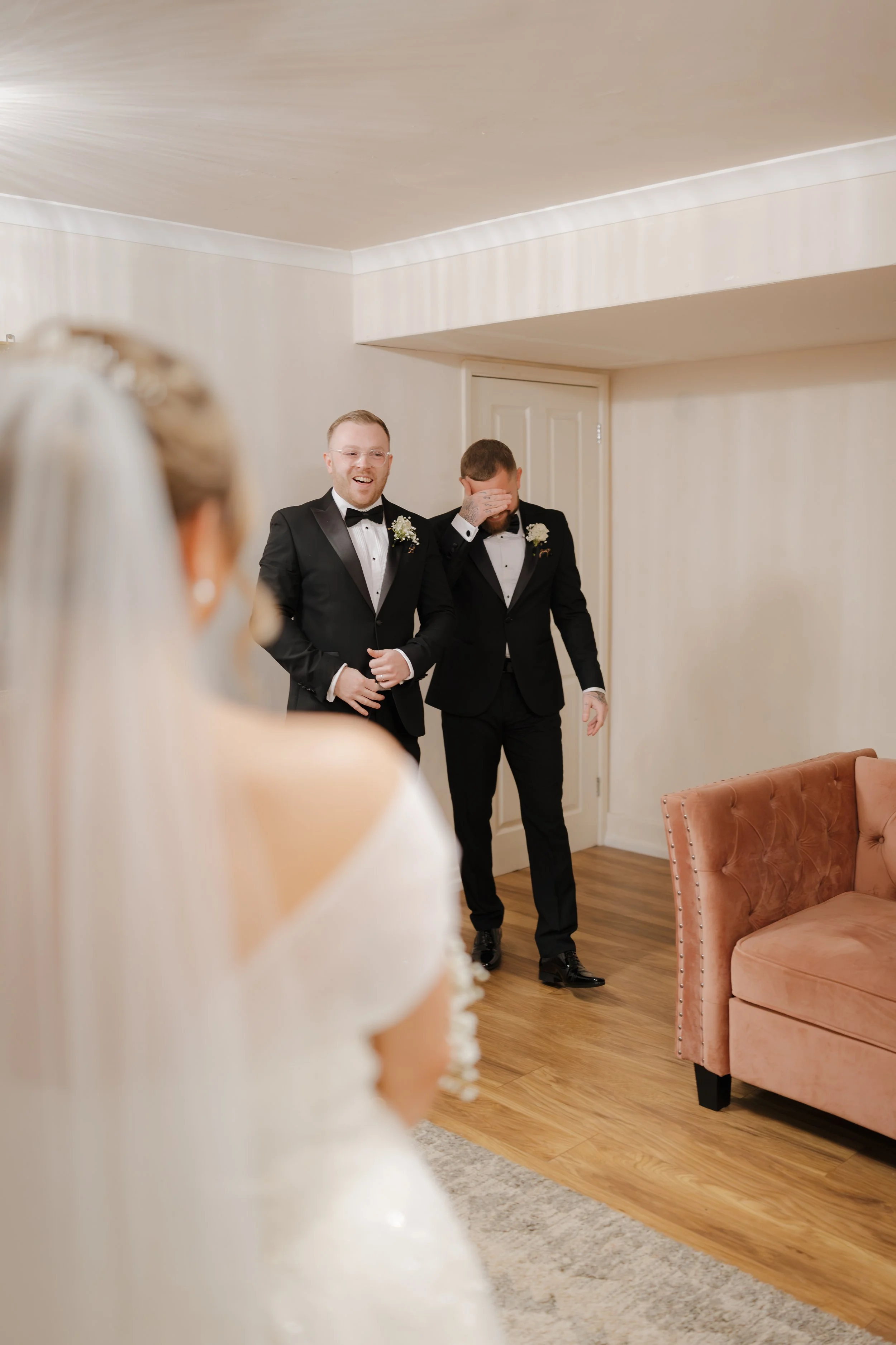 Two men dressed in black tuxedos smile and laugh, standing in a room with beige walls, as a woman in white wedding dress watches them, her back to the camera.
