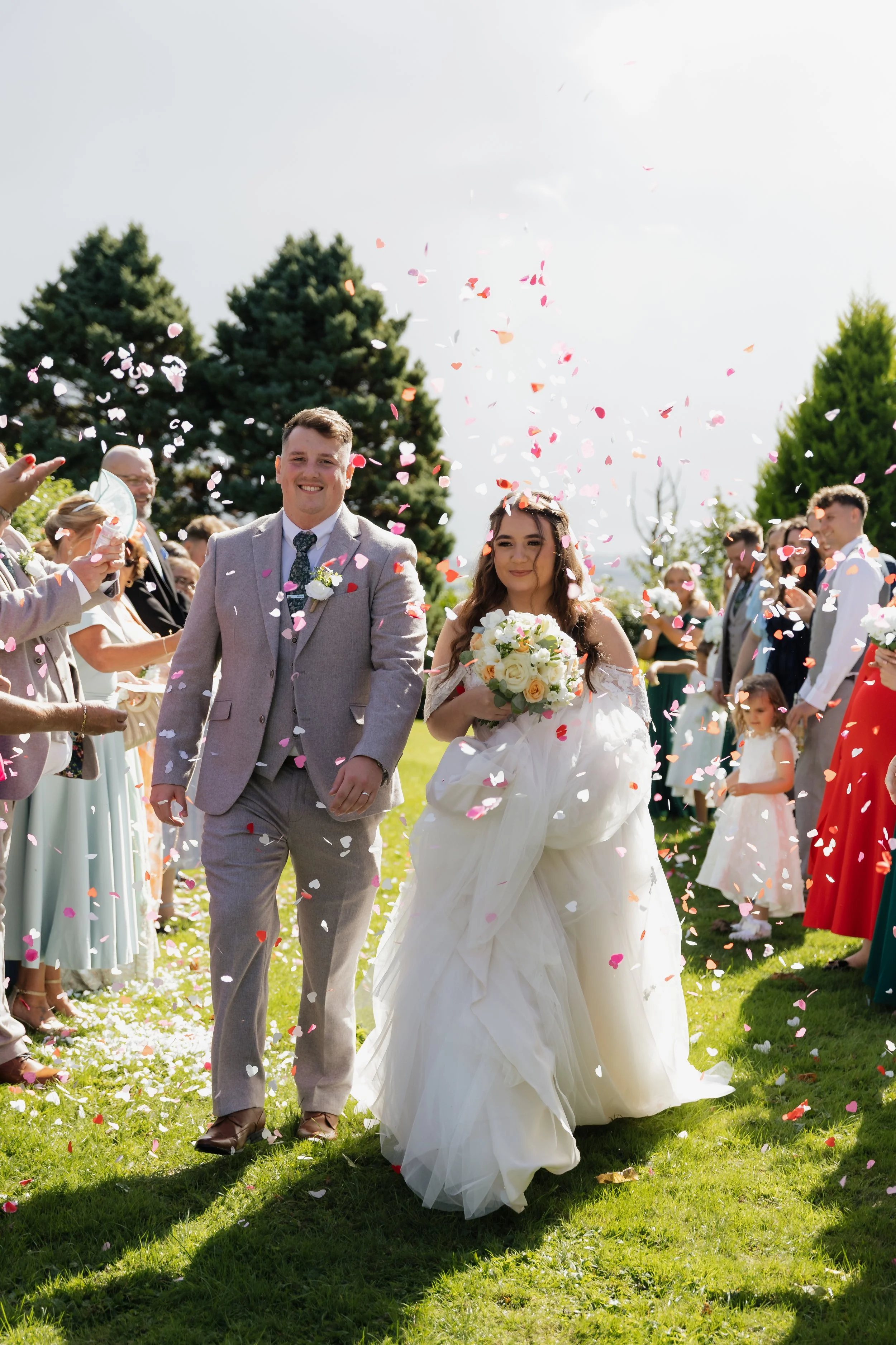 Close-up of a bride in a lace wedding dress holding a bouquet of white roses. Hands with wedding rings are placed on her shoulder, revealing a bracelet and elegant fingernails.