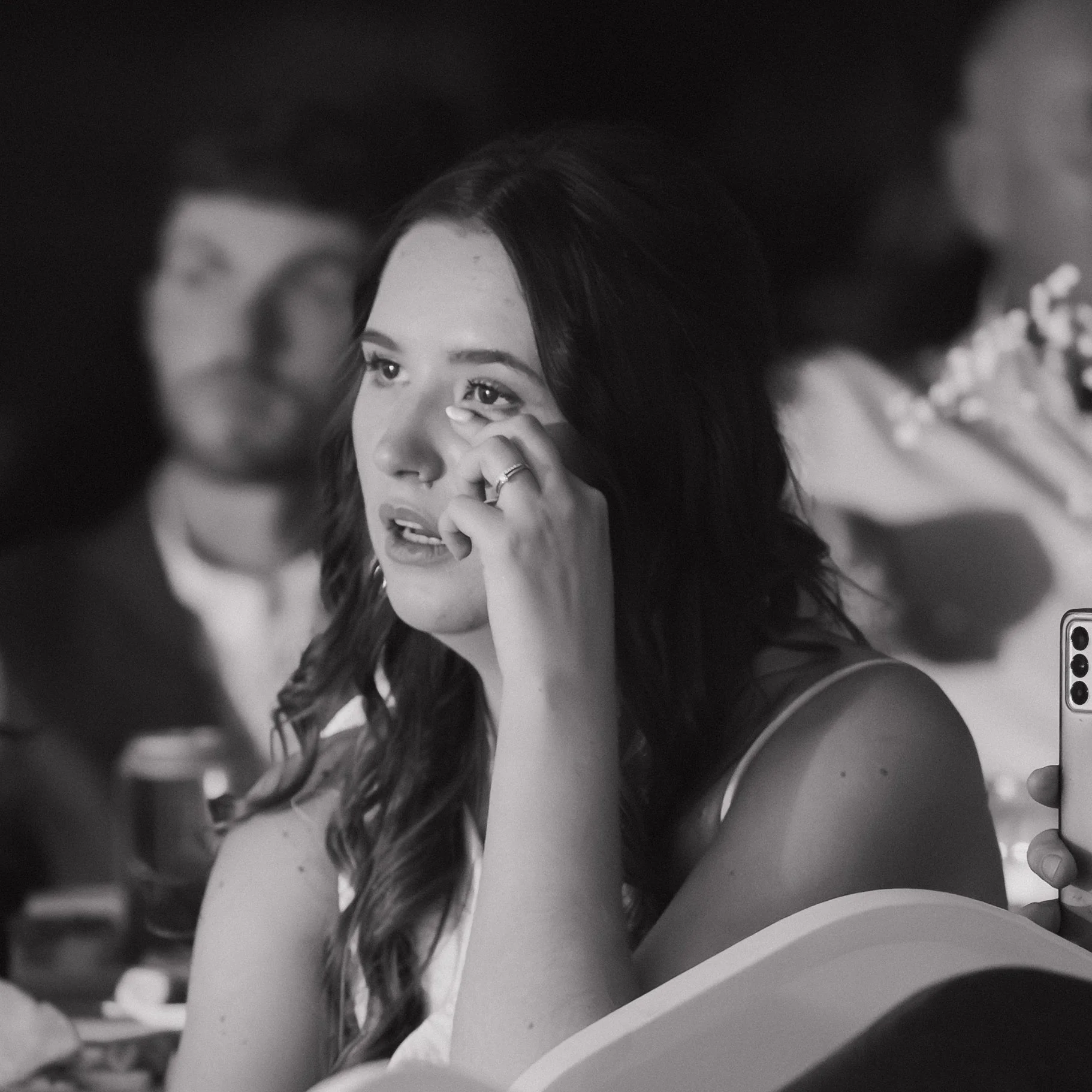 Black and white photo of a woman with long, wavy hair touching her face, appearing emotional, with another person partially visible in the foreground holding a phone.