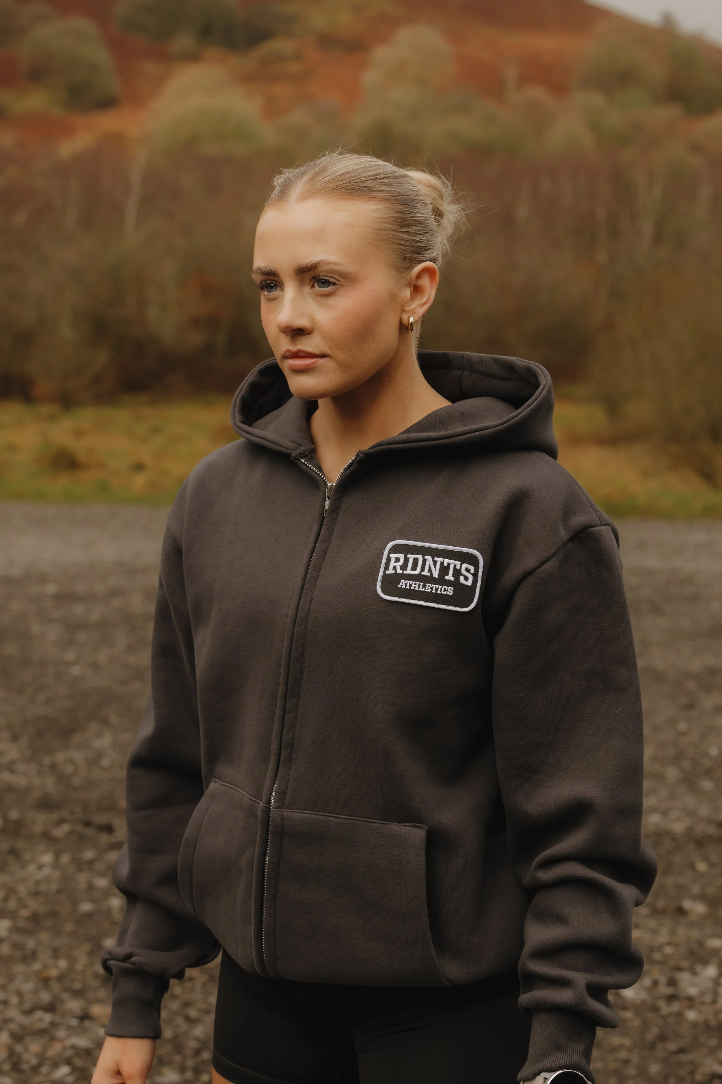 A woman standing outdoors on a gravel surface with autumn trees in the background, wearing a dark hoodie with an 'RDNT'S ATHLETICS' patch on the chest.
