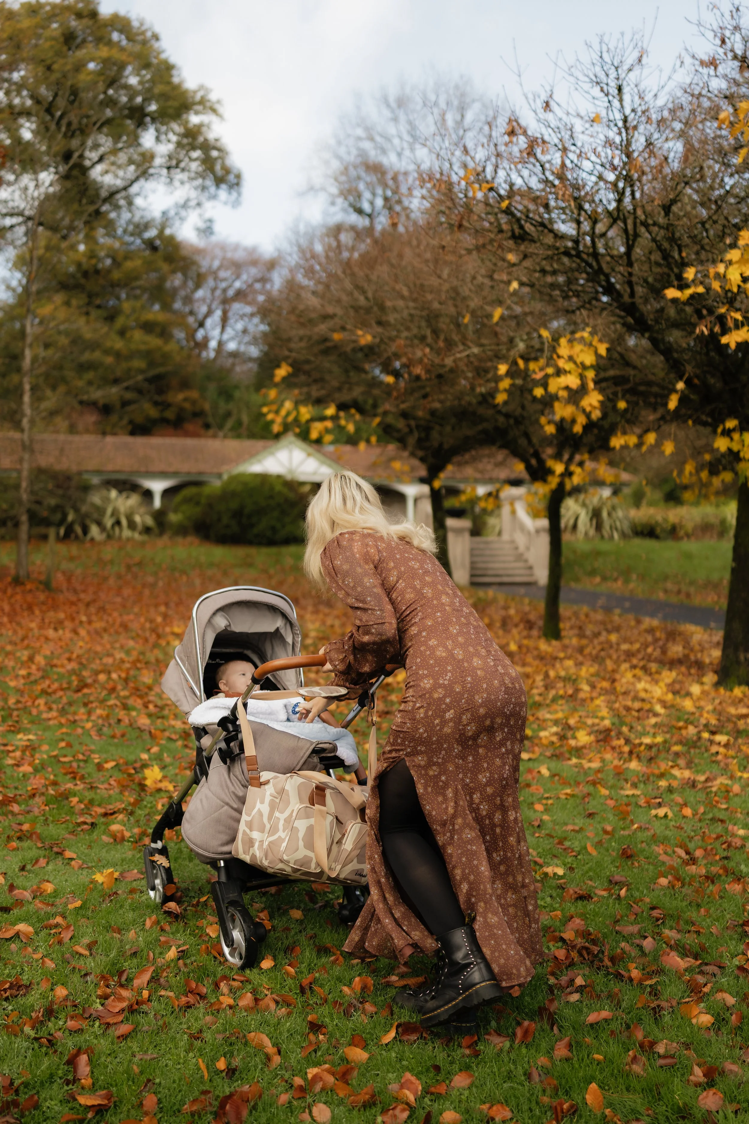 A woman leaning over a stroller with a baby inside in a park during autumn, with fallen leaves on the ground and trees with mostly bare branches.