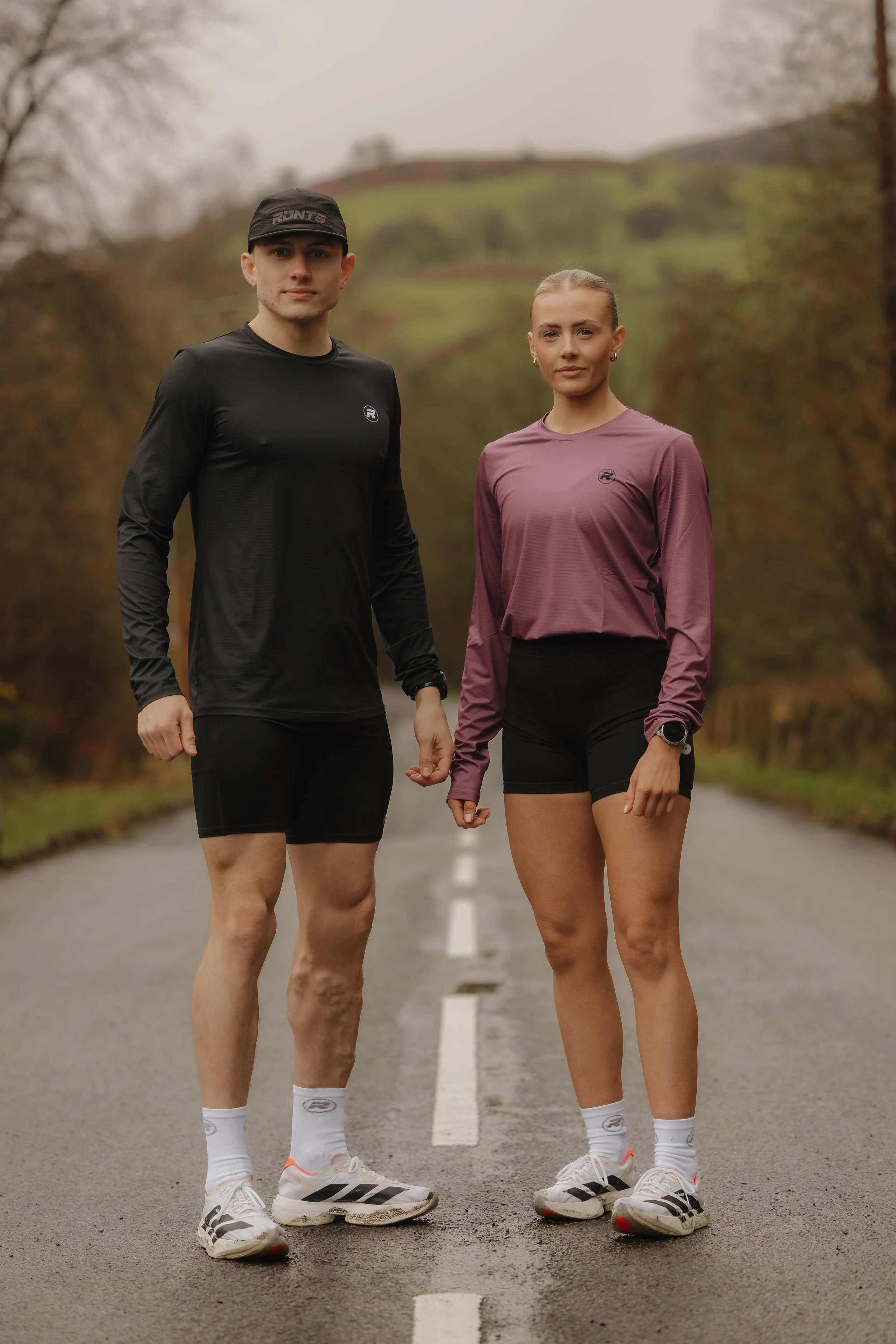 A man and a woman dressed in athletic wear, standing hand in hand on a rural road with a hilly, wooded landscape in the background.