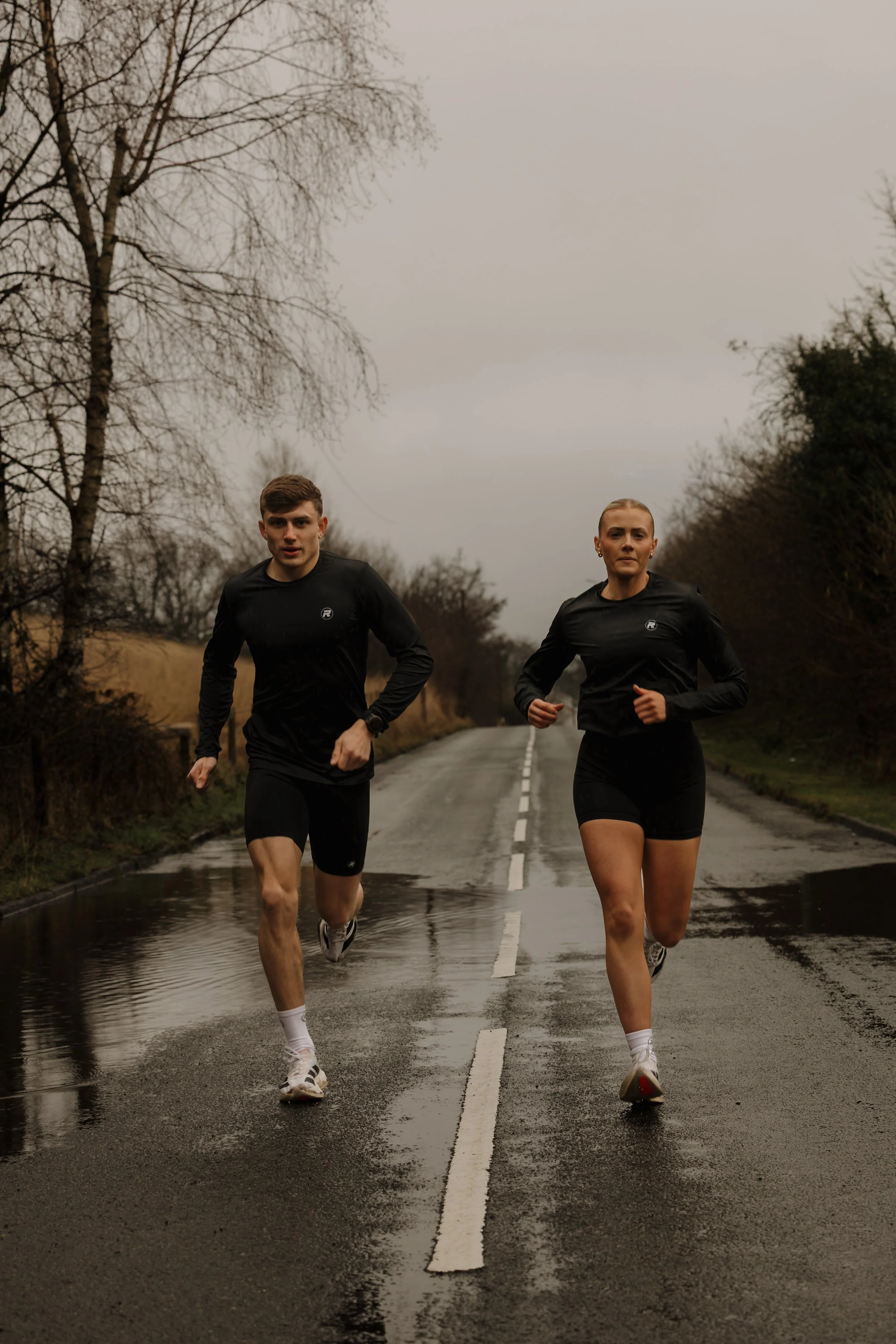 A man and a woman jogging on a wet, empty road during overcast weather with leafless trees on both sides.