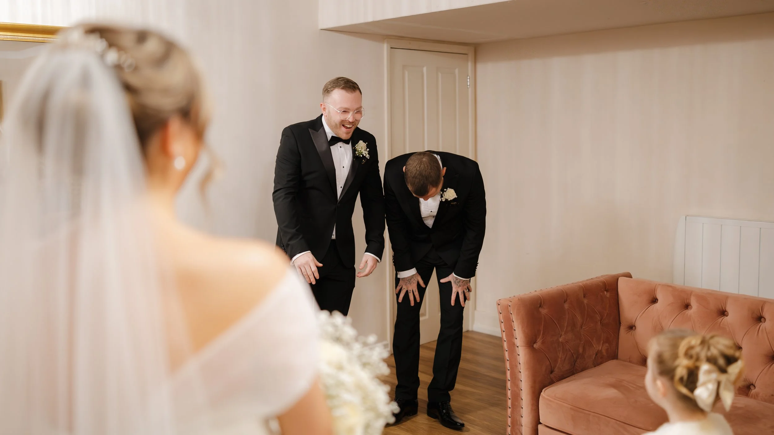 Two men in black tuxedos laughing and bowing inside a room, with a bride and a girl in the foreground.