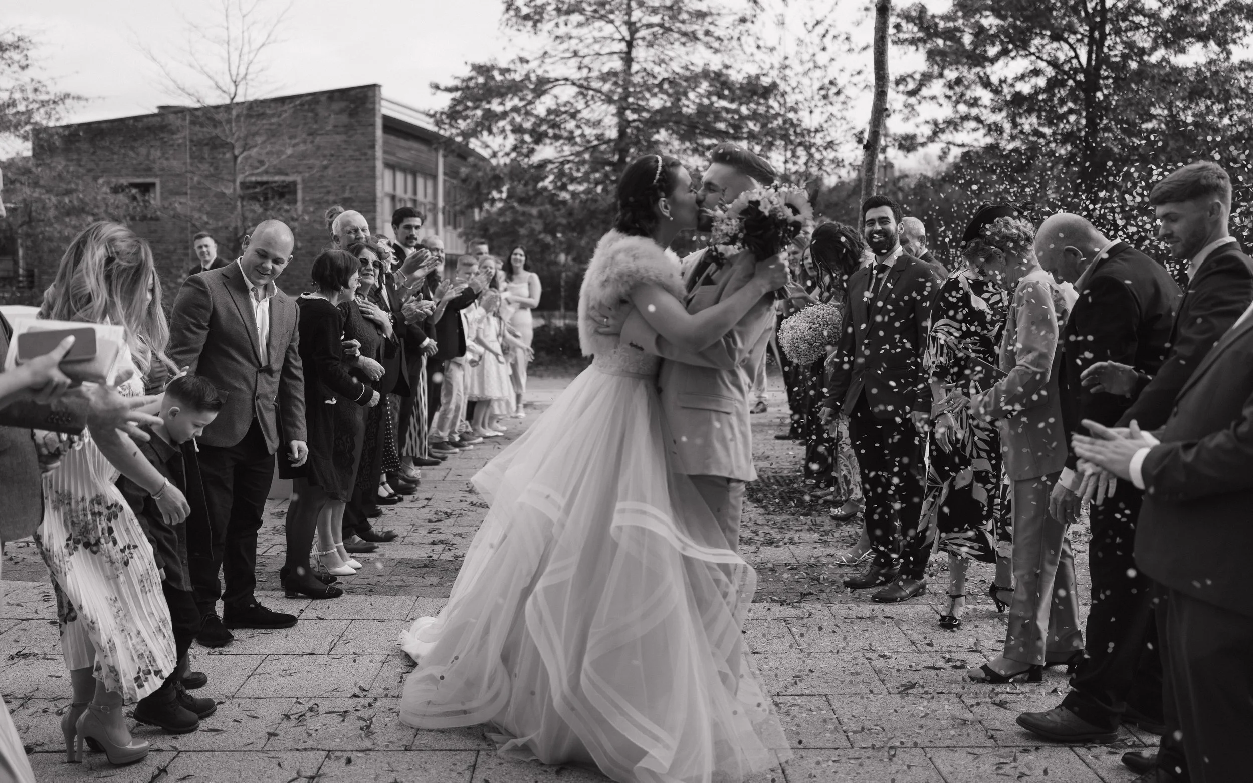 A newlywed couple shares a kiss and hugs in the middle of an outdoor wedding celebration as guests cheer and clap around them.