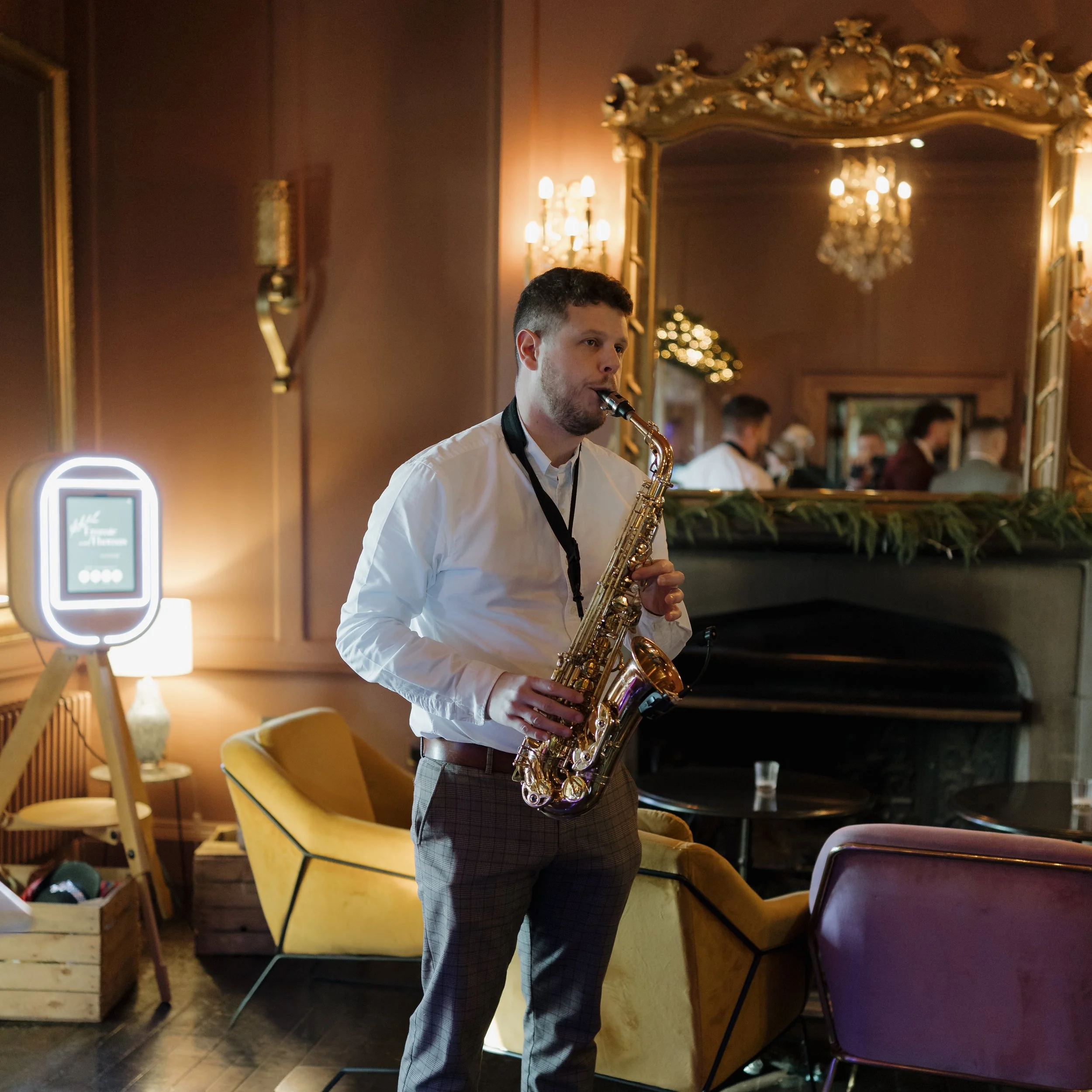 A man playing a saxophone at an indoor event with elegant décor, including ornate mirrors, chandeliers, and cozy seating.