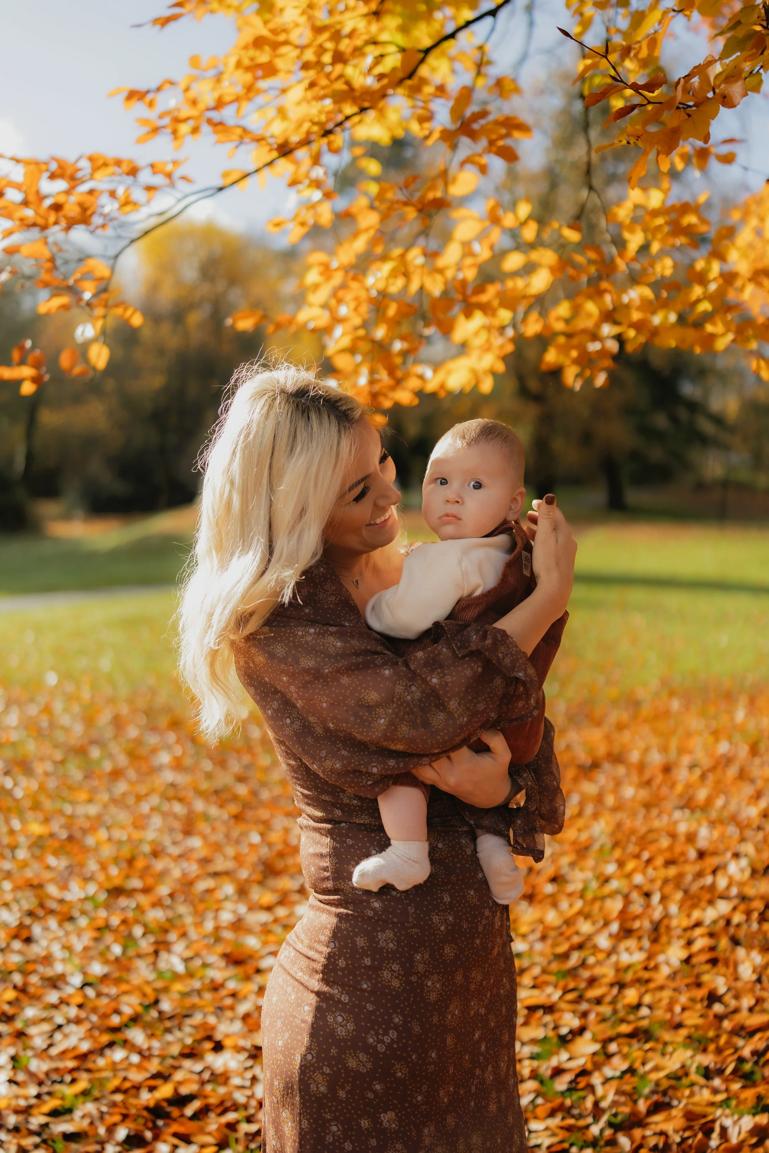 A woman with blonde hair, smiling, holding a baby in an outdoor park during autumn with fallen leaves and orange trees.