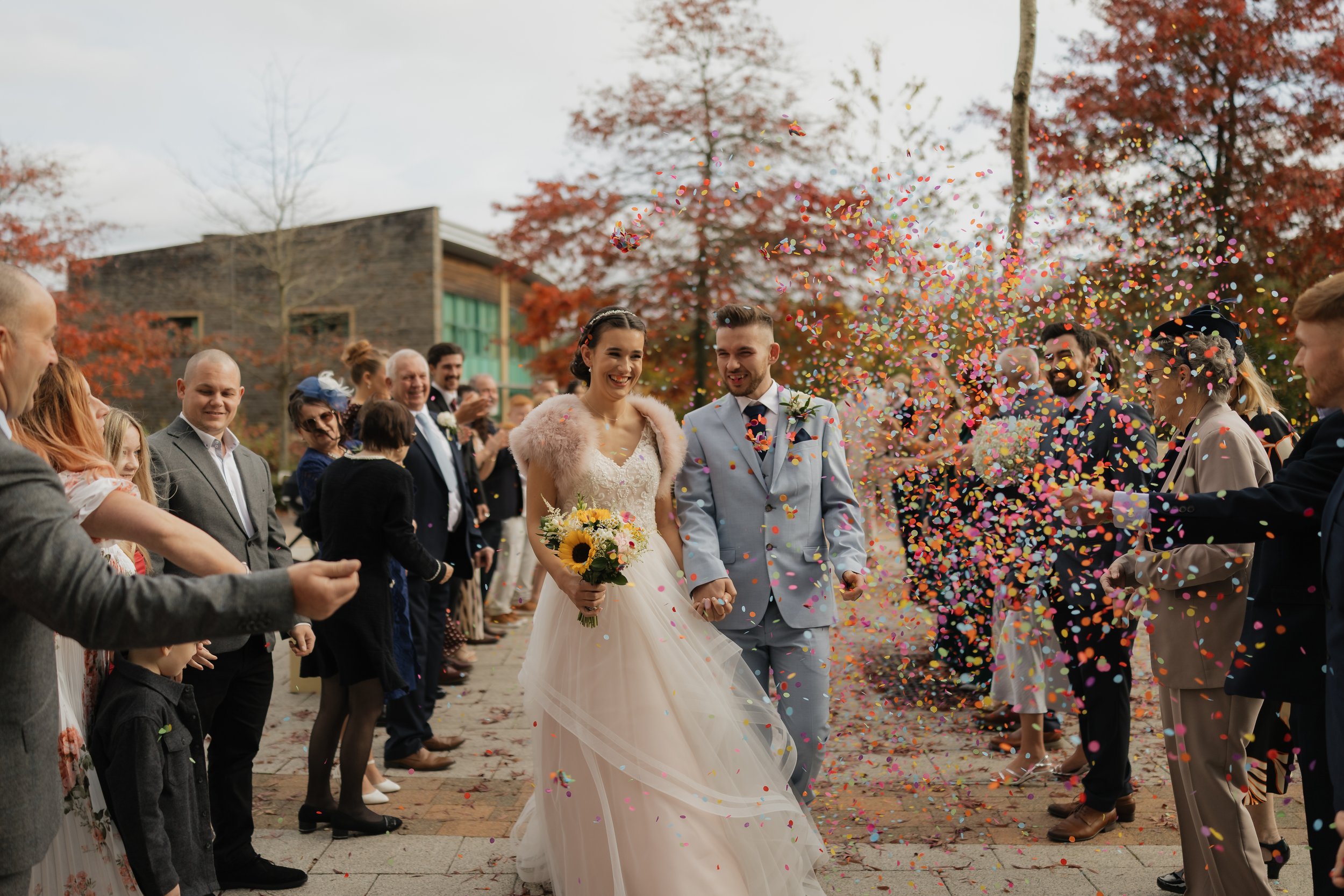 A bride and groom smiling and holding hands as they walk through a crowd of guests throwing confetti during their wedding celebration outside, with fall-colored trees and modern building in the background.