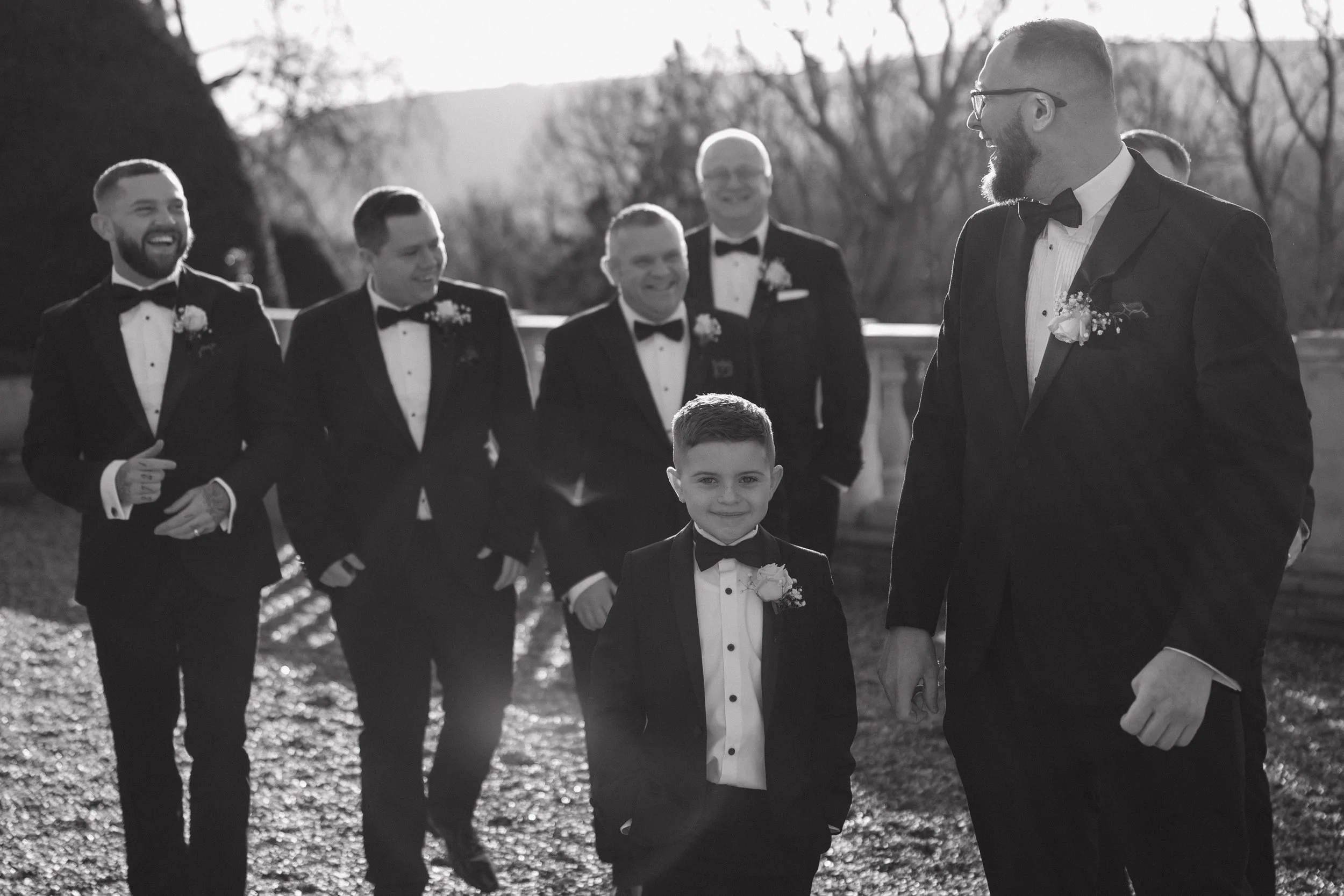 A group of men and a young boy dressed in tuxedos, standing outdoors, smiling and laughing in a social gathering or wedding event.
